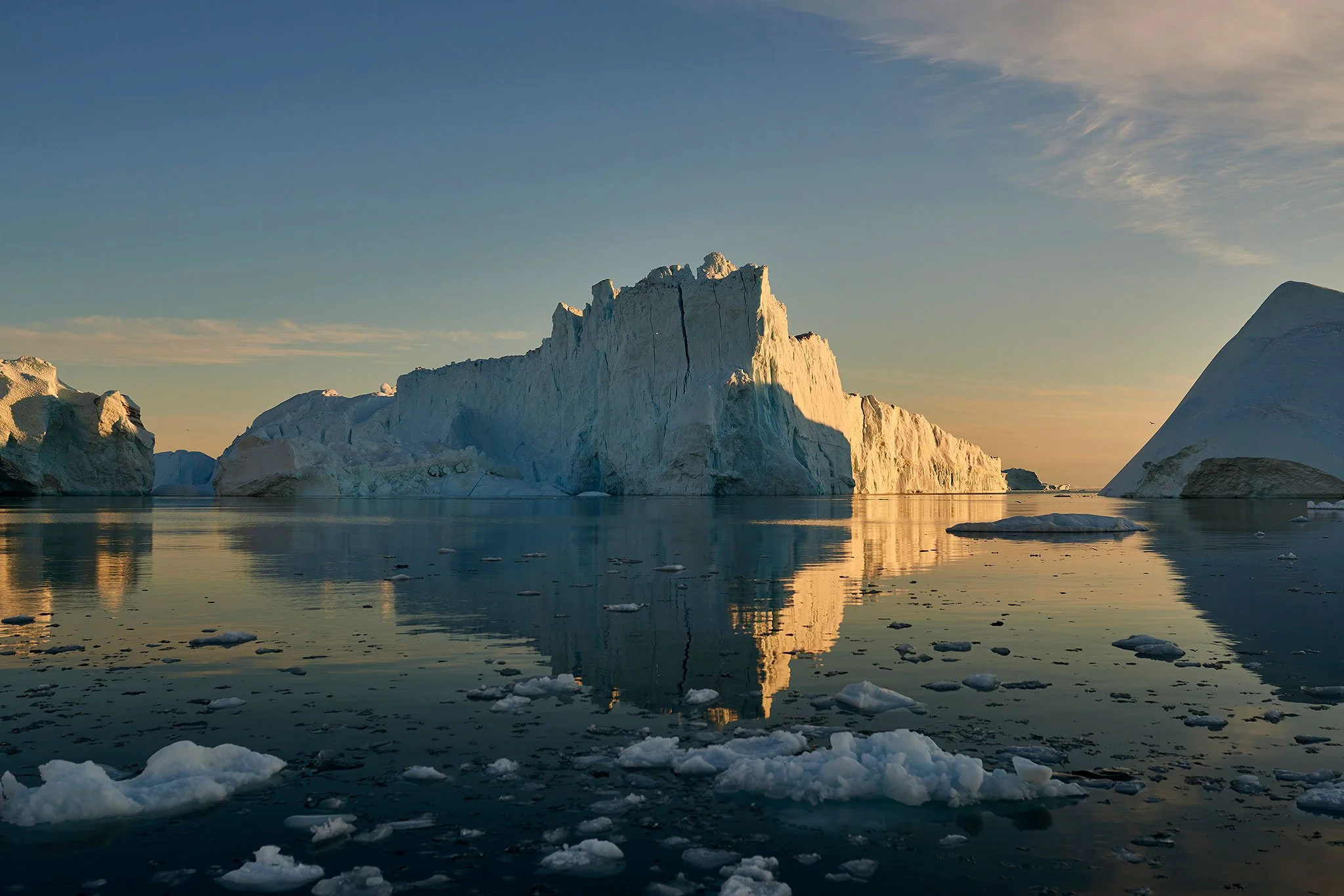Atmospheric iceberg landscape with soft dawn light, icy detail and mirror-like reflections across a tranquil Arctic seascape.
