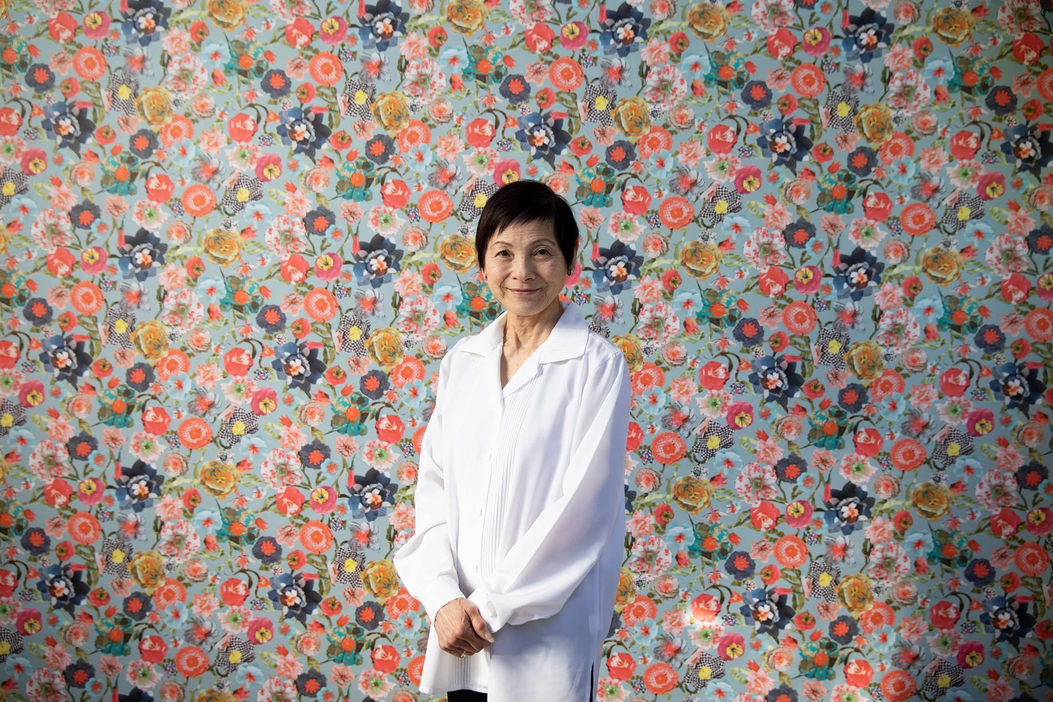 Portrait of a person in a white robe standing before a colorful floral wall.