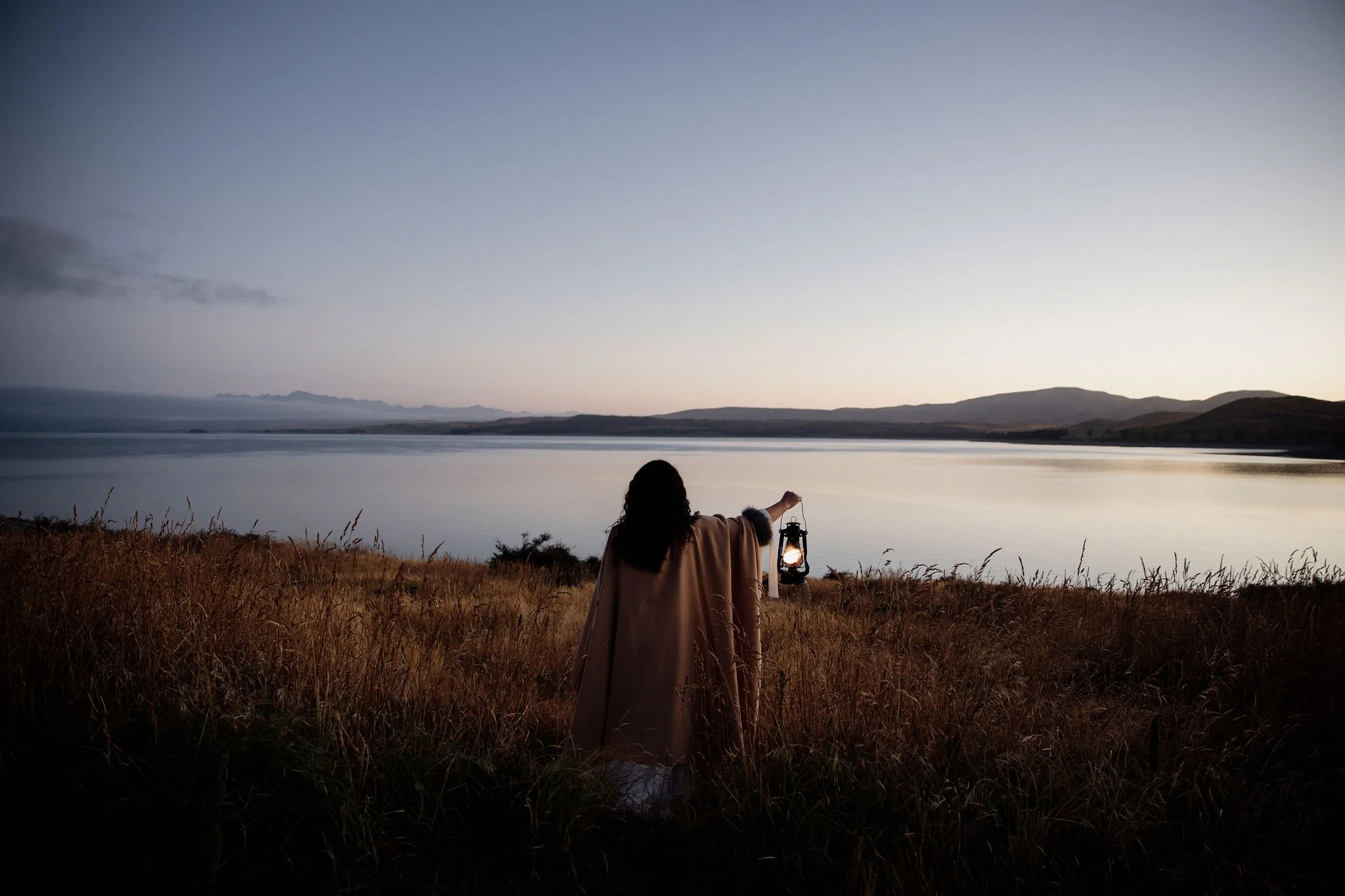 Woman standing beside a quiet lake at dusk with soft horizon light behind her.