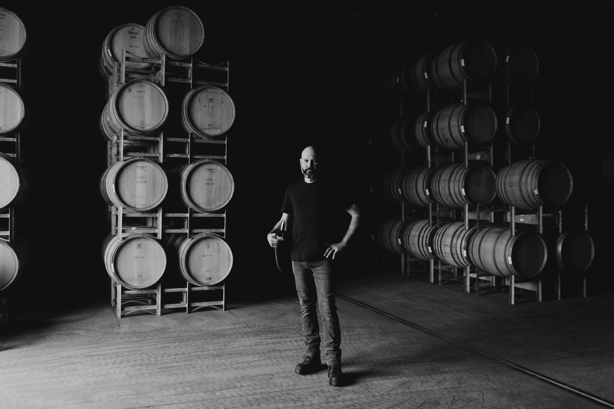 Black-and-white portrait of a winemaker standing among stacked barrels.