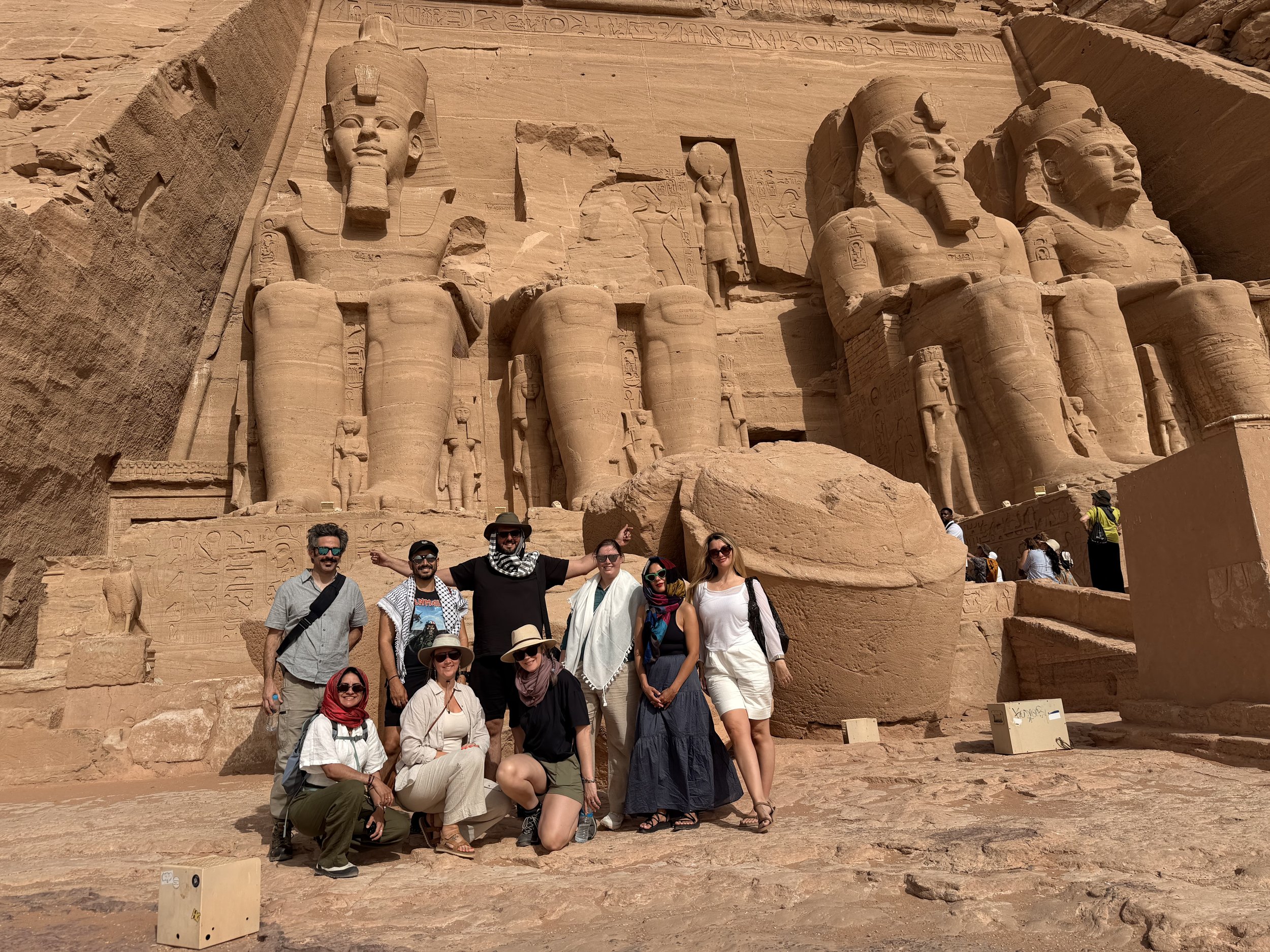 A group of tourists standing and kneeling in front of the Great Pyramid of Giza, with large carved Egyptian statues and hieroglyphics on the pyramid wall behind them.