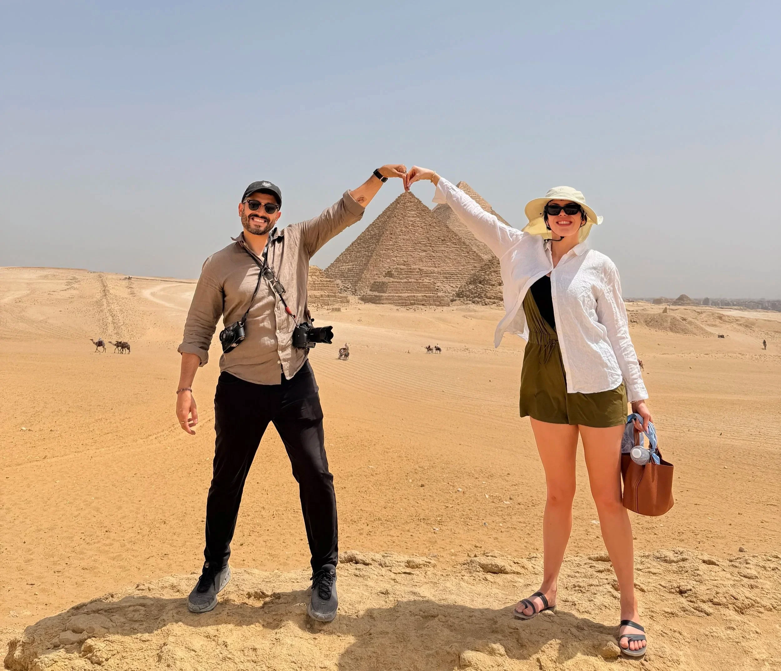 Two tourists making heart shapes with their hands above the pyramids in the desert with camels in the background.