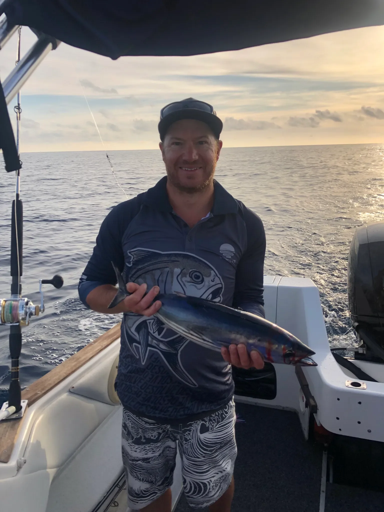 A man holding a fish on a boat with the ocean and sunset in the background.
