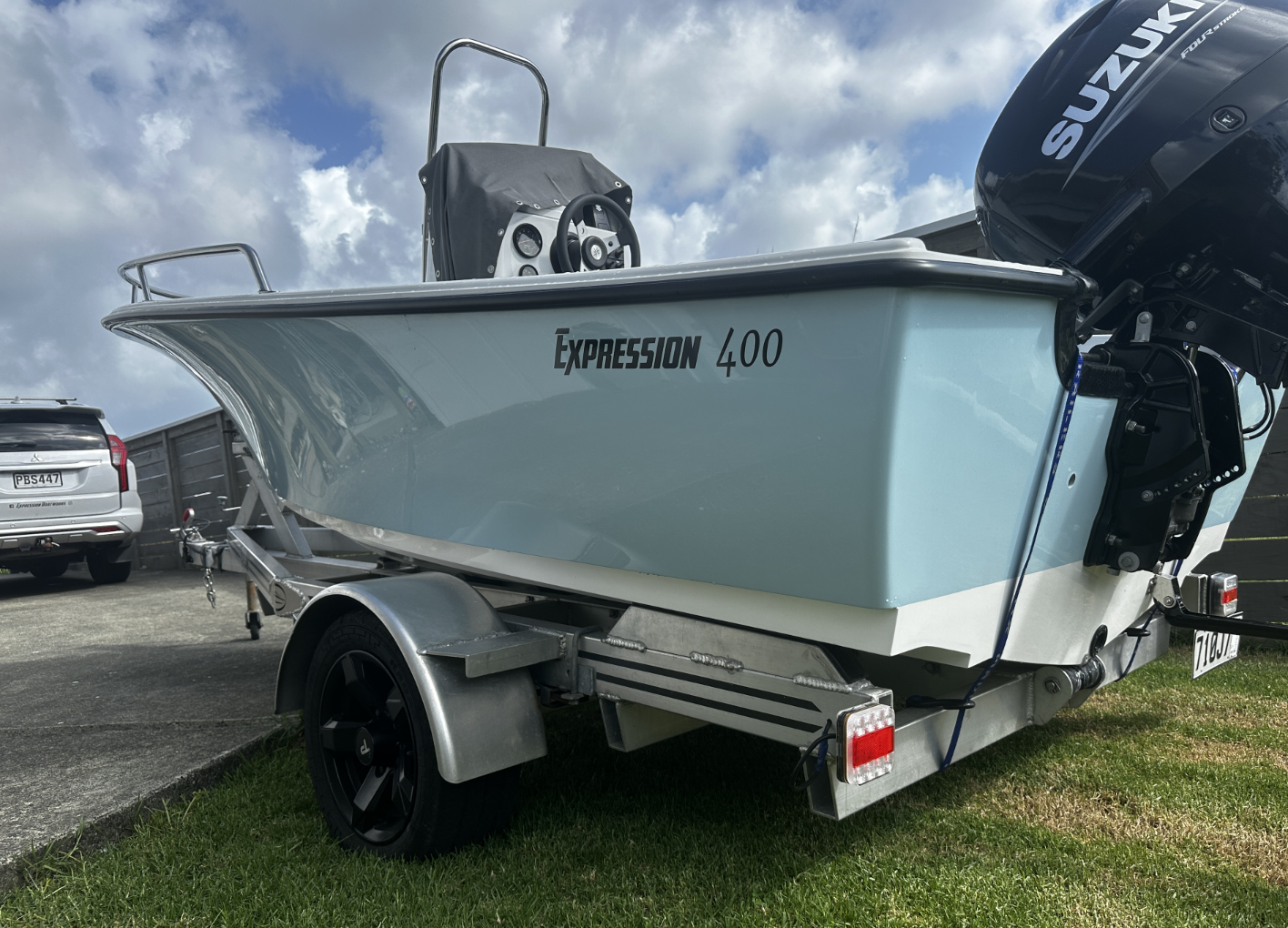 Light blue boat on a trailer with a Suzuki outboard motor, parked on grass and pavement, under cloudy sky.