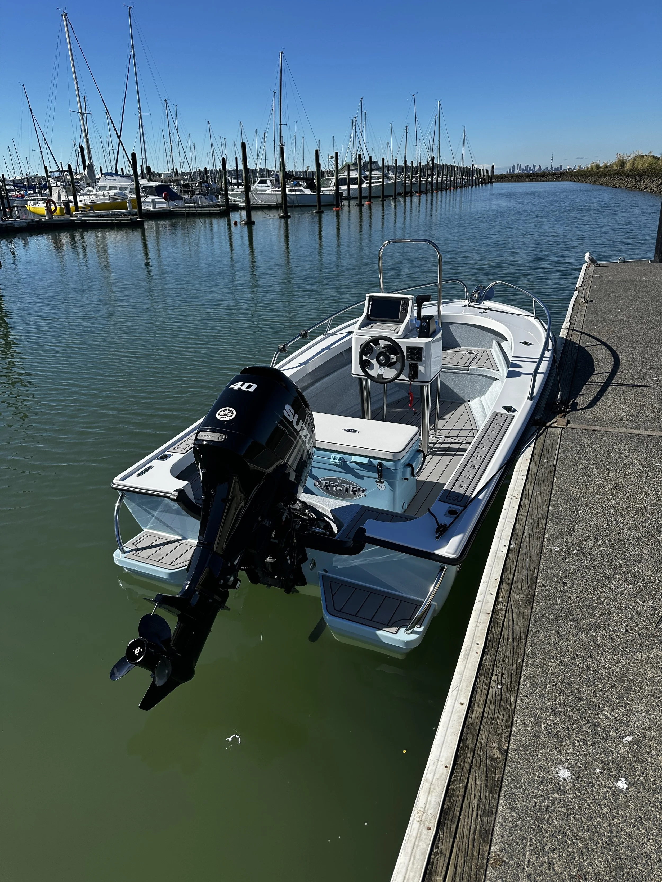 A small white motorboat docked at a wooden pier in a marina, with multiple sailboats and yachts moored in the background under a clear blue sky.