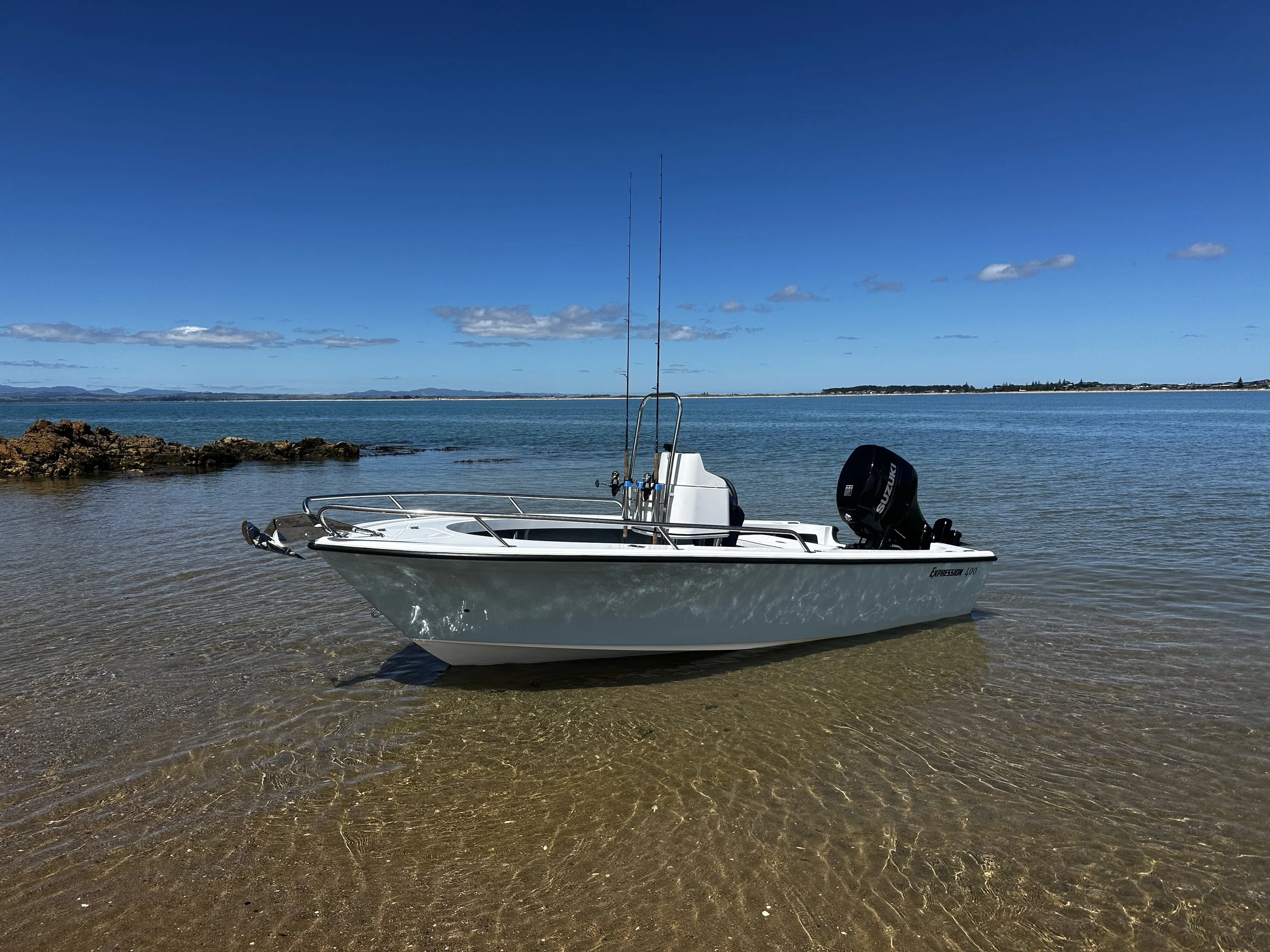 A small white boat with a Suzuki engine floating near the shore on clear water, with distant land and blue sky with scattered clouds in the background.