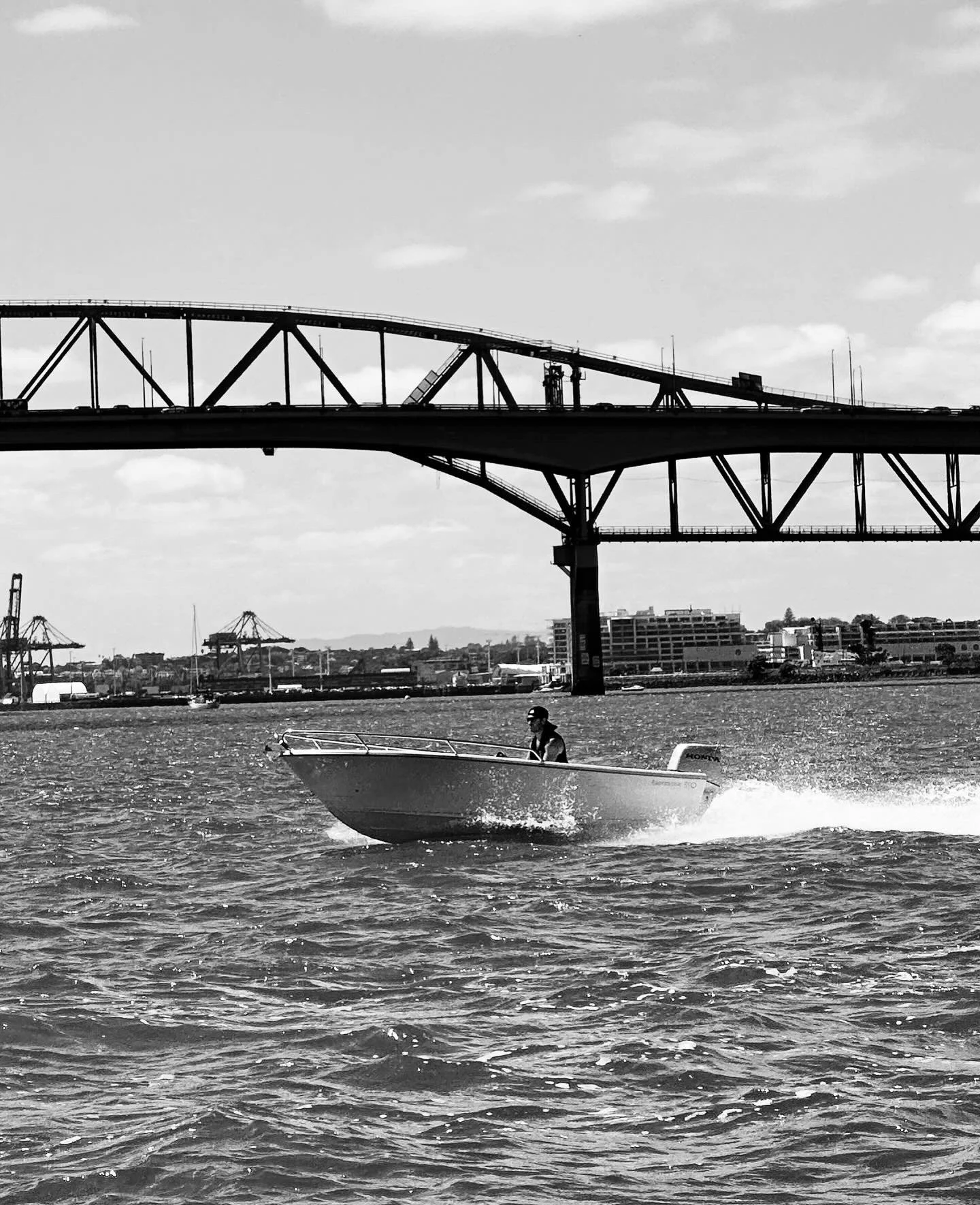 A person wearing a hat or helmet is riding a motorboat on a body of water, with large bridges and industrial cranes in the background.