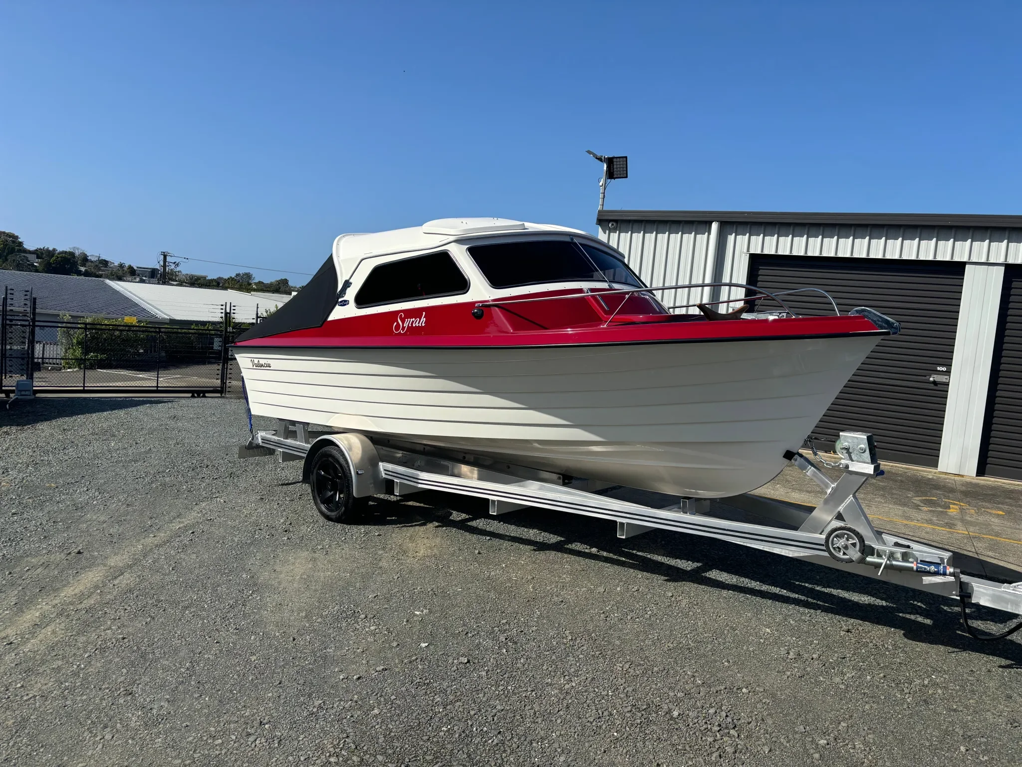A small motorboat with a red and white hull named 'Syrah' on a trailer in a parking lot, with a building and clear blue sky in the background.