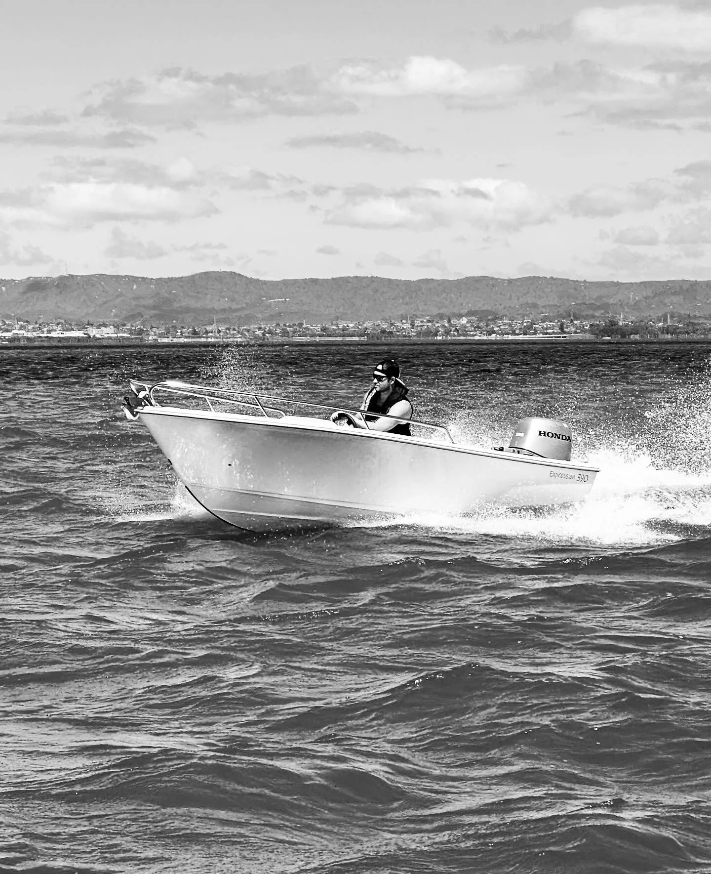 A person in a small motorboat on a body of water with a mountain range in the background and partly cloudy sky