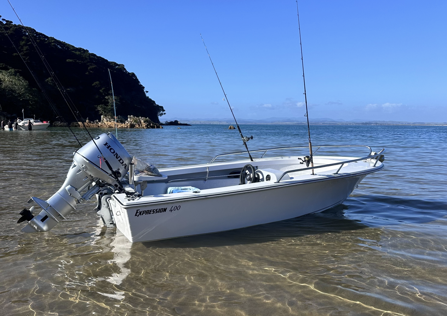 A small white boat with a Honda outboard motor in shallow water near a rocky shoreline with trees, under a clear blue sky.