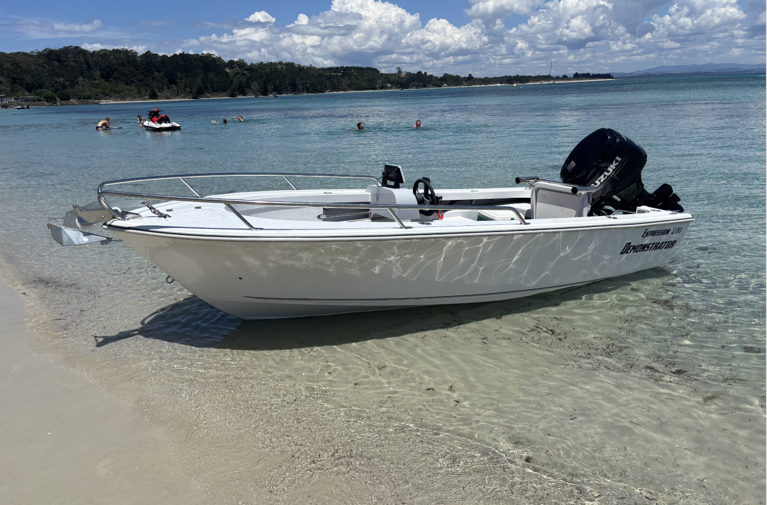 A small white boat with a Suzuki outboard motor, anchored near the shore of a beach with clear shallow water. In the background, a few people are swimming and floating in the water, with a lush green hillside and a partly cloudy sky overhead.