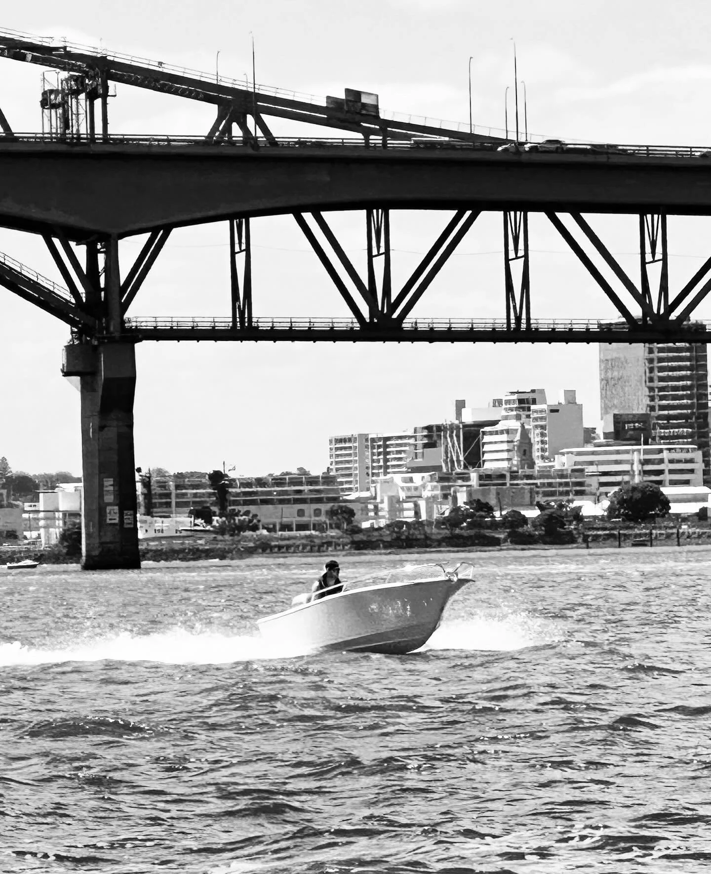 A black and white photo of a boat on a river with a city skyline and a large bridge overhead.