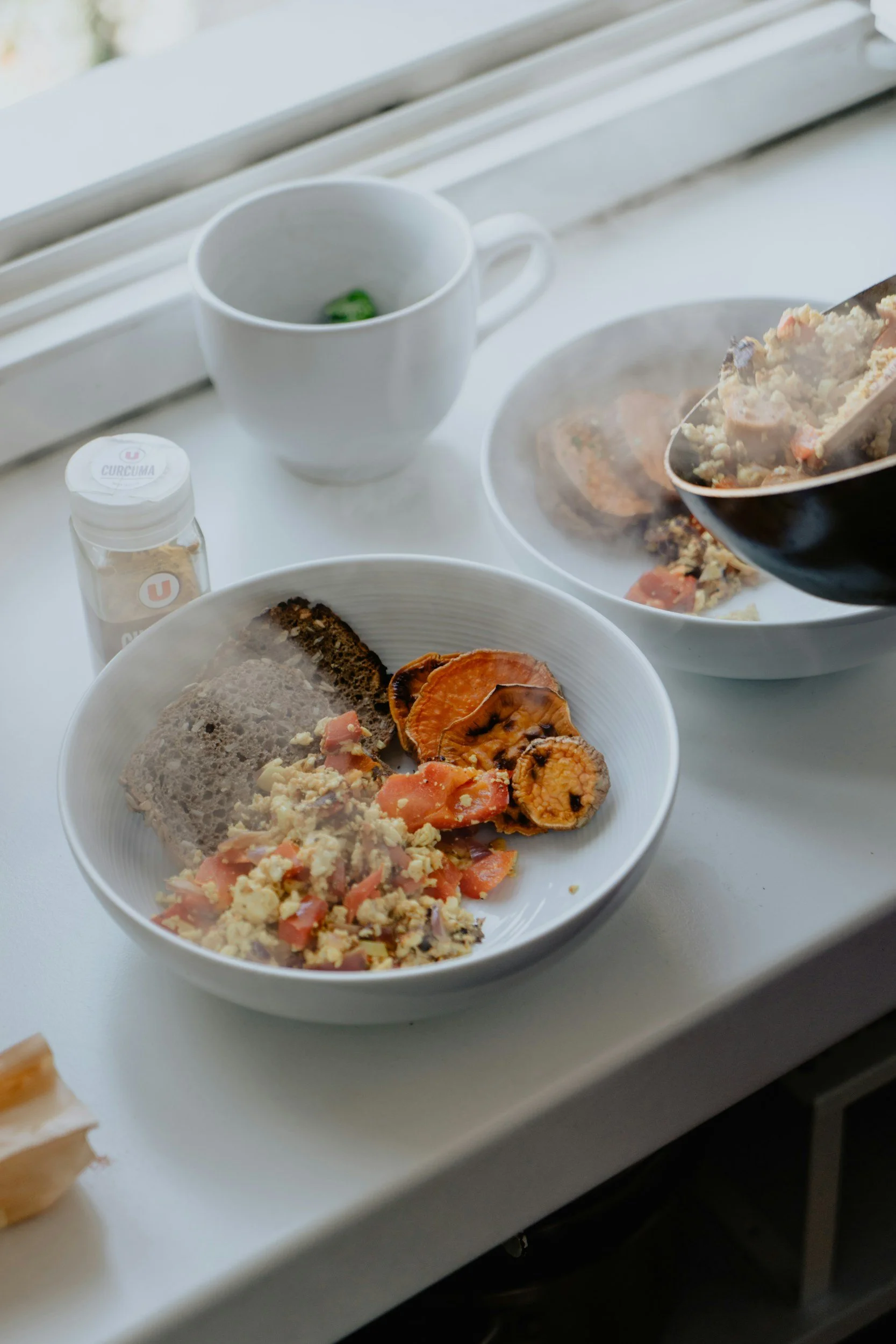 A white bowl with scrambled eggs, sliced smoked salmon, and cooked vegetables. A white cup with a green object inside. A container labeled 'Curcuma' on a white surface near a window.