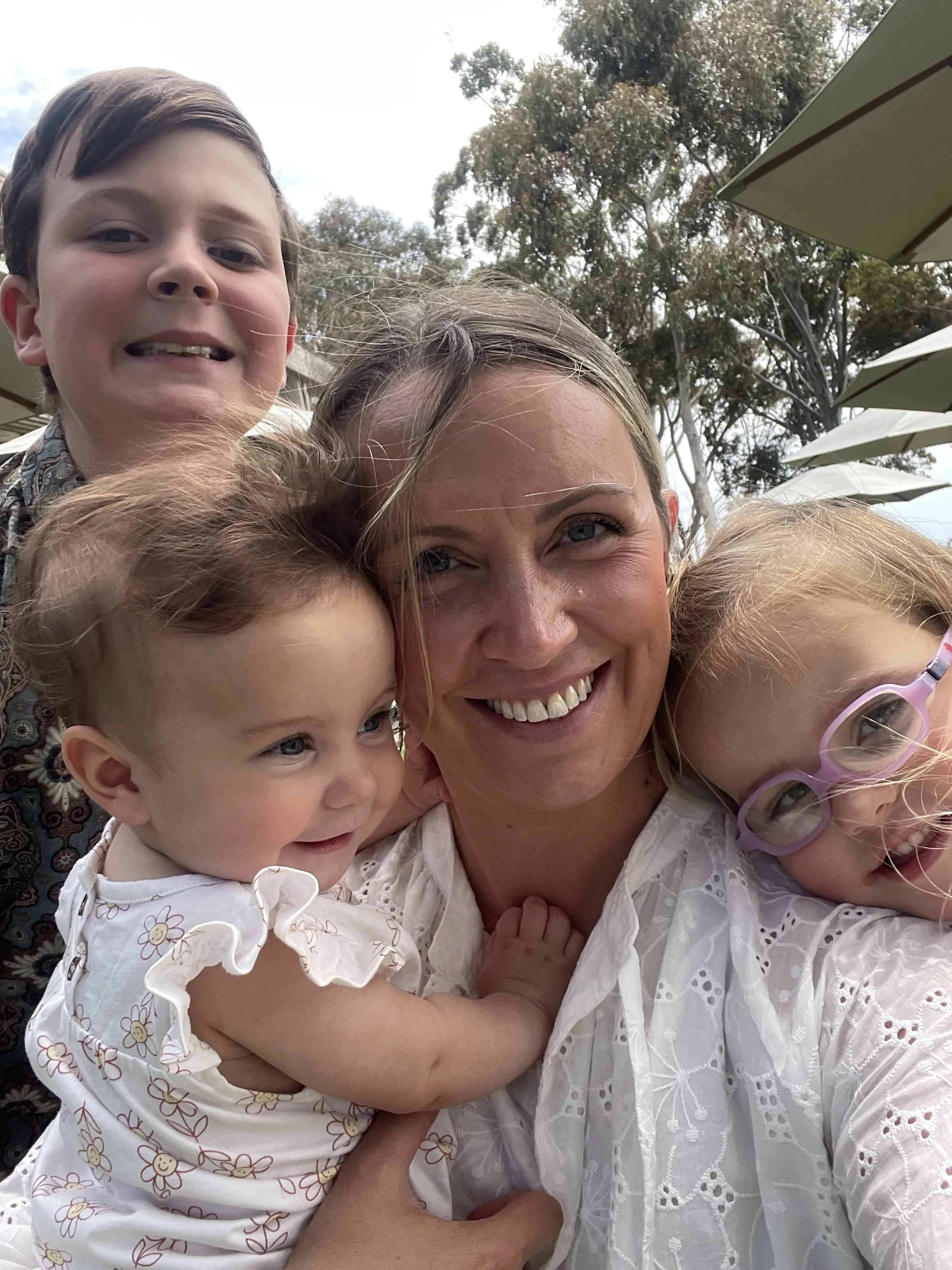 Smiling woman with blonde hair taking a selfie with four young children outdoors. The children have light hair and are wearing white clothing with floral patterns, and one girl has pink glasses. There are trees and umbrellas in the background.