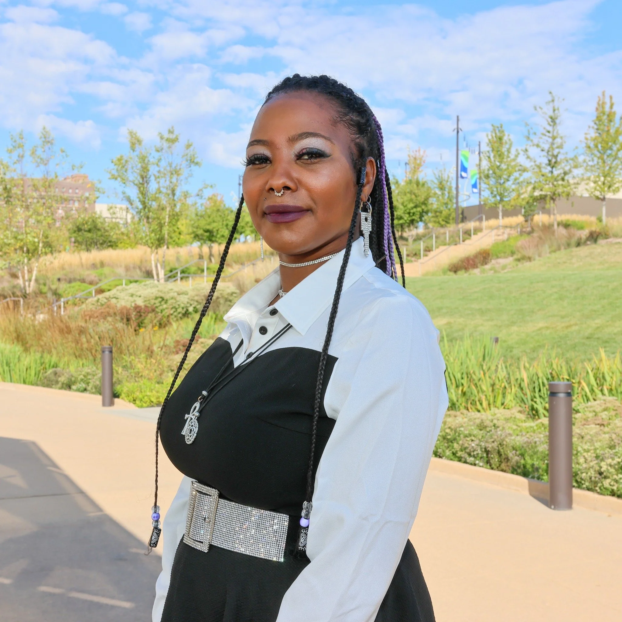 A close-up photograph of a woman with braided hair wearing earrings and a nose ring, standing outdoors against a background of trees, a pathway, and colorful banners, under a partly cloudy sky.