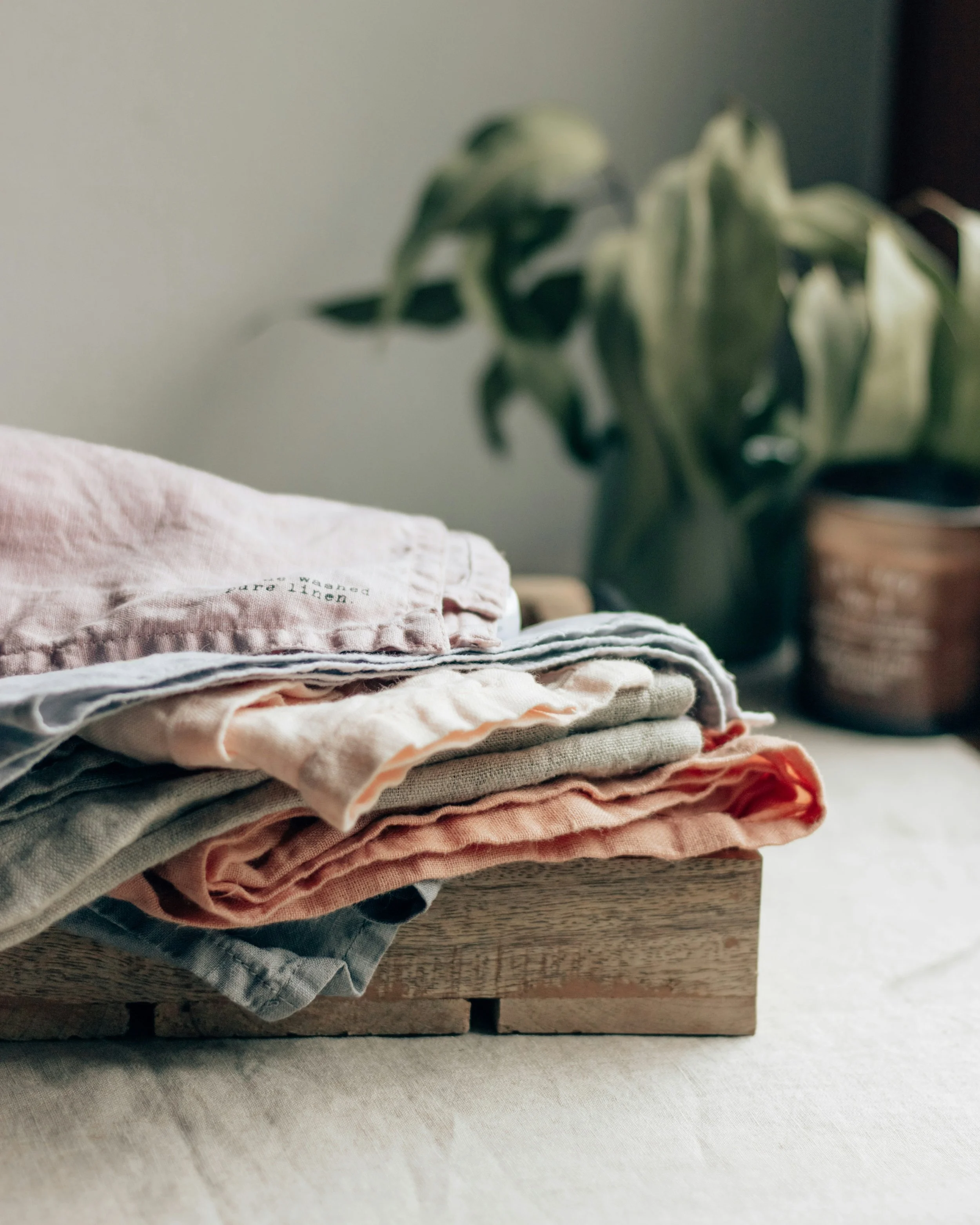 Stacked folded clothes on a wooden surface with a blurred plant in the background.