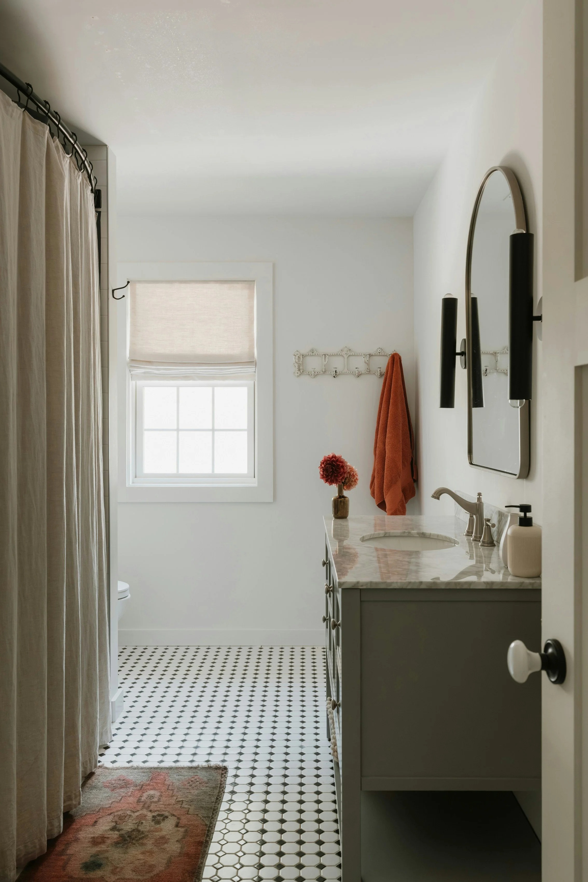 A cozy bathroom with a black and white hexagon tile floor, a white vanity with marble countertop, a small vase with red flowers, a towel hanging on a white rack, a mirror with black wall sconces, a window with a beige Roman shade, and a shower curtain on the left.