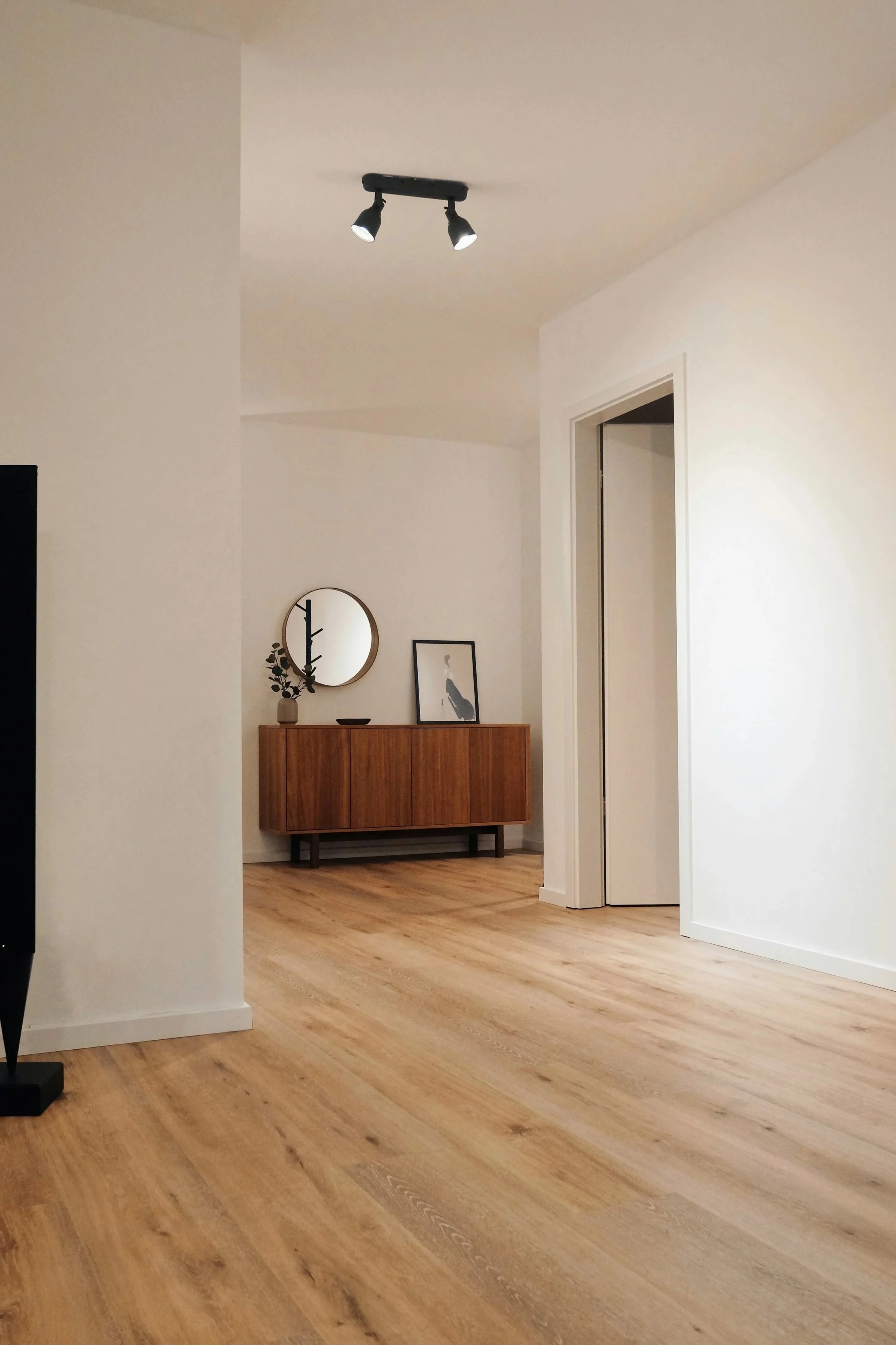 Living room corner with off-white walls, a wooden sideboard, a round mirror, framed artwork, and a ceiling light fixture with two adjustable spotlights.