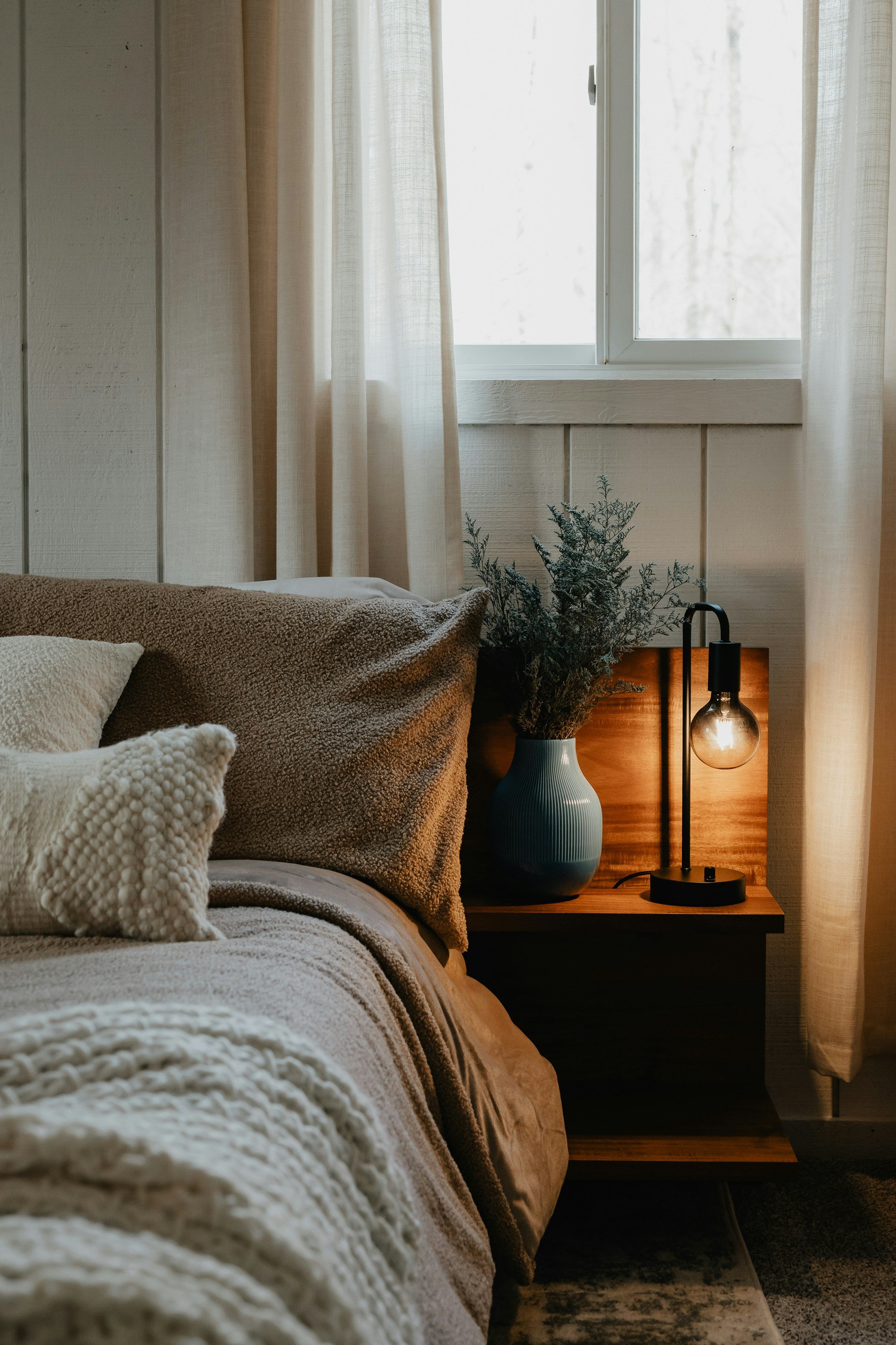 A cozy bedroom with a bed, beige and textured pillows, a comforter, a large window with white curtains, a potted plant in a white vase near the bed, and a black table lamp with an exposed bulb on a wooden nightstand.