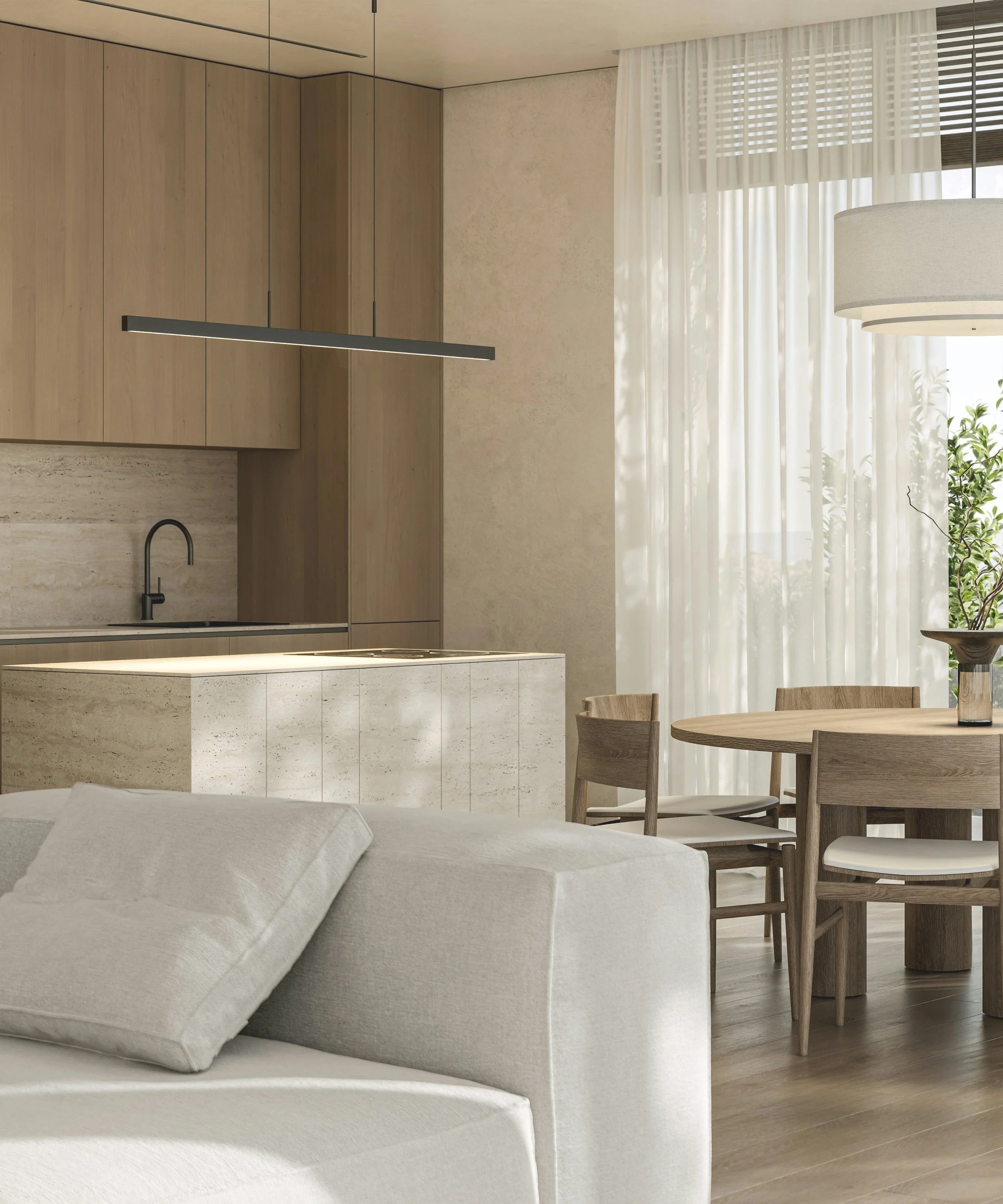 A modern, minimalist kitchen and dining area with light wood cabinets, a round wooden dining table with matching chairs, sheer white curtains, and a large beige upholstered sofa.