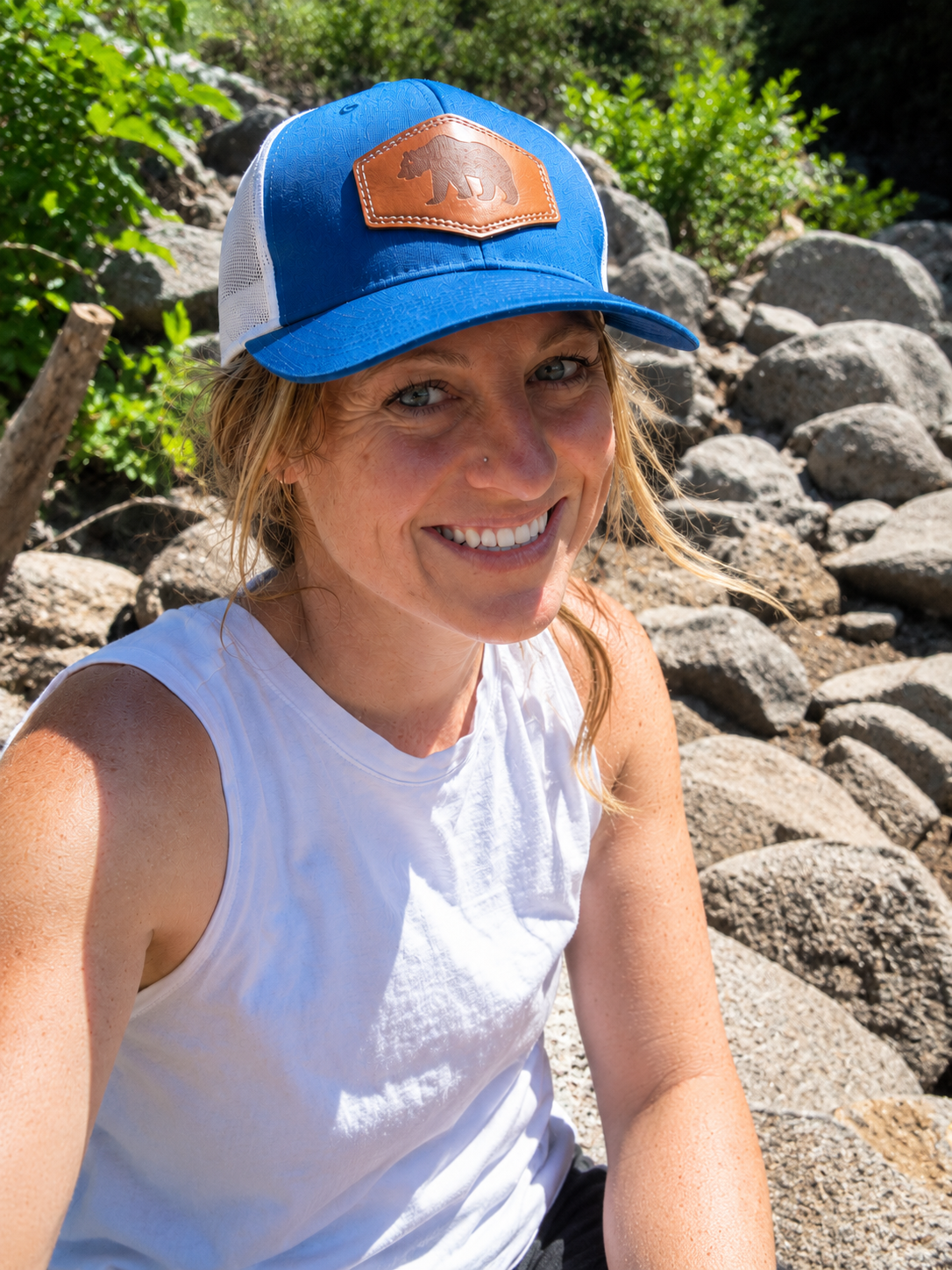 A smiling woman wearing a white sleeveless shirt and a blue trucker hat with a tan bear patch, sitting on rocks outdoors with greenery in the background.