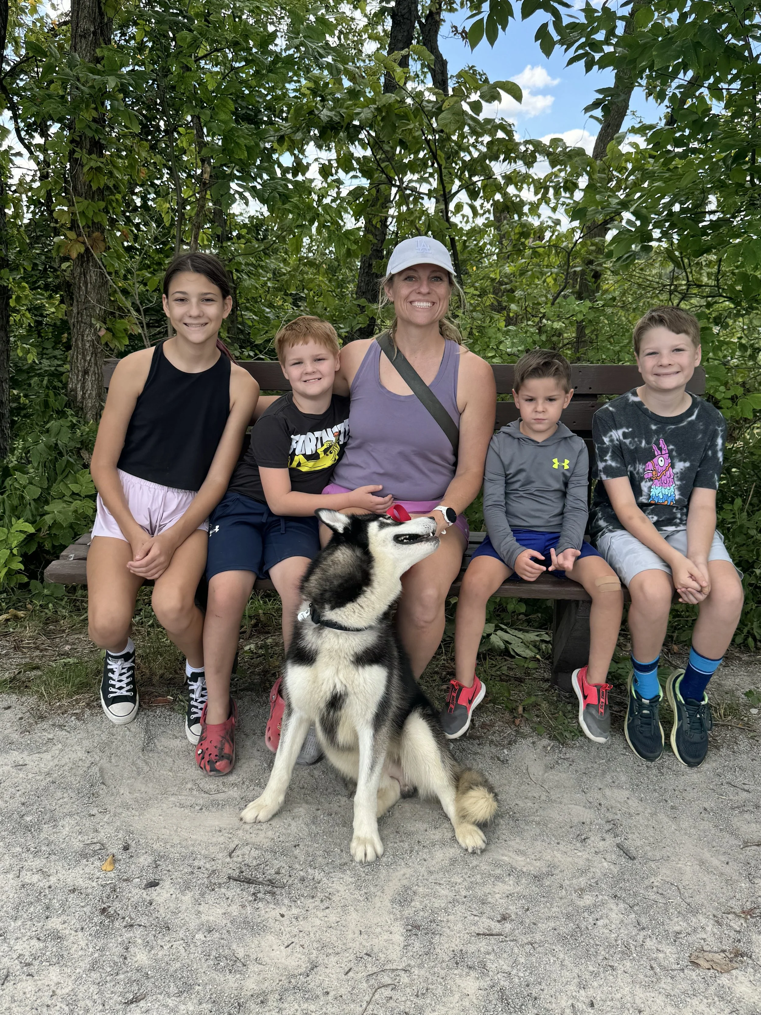 A woman and five children sitting on a park bench outdoors, with a husky dog sitting on the ground in front of them. The background is filled with green trees and blue sky.