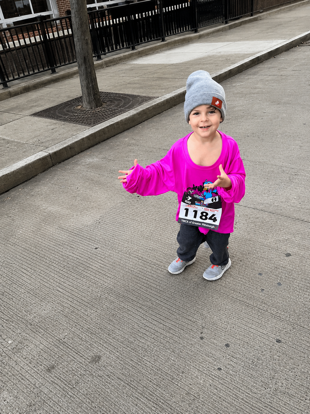 Young child smiling and pointing, wearing a gray beanie, pink long sleeve shirt with a race bib labeled 1184, black pants, and gray sneakers, standing on a sidewalk near a tree and black railing.