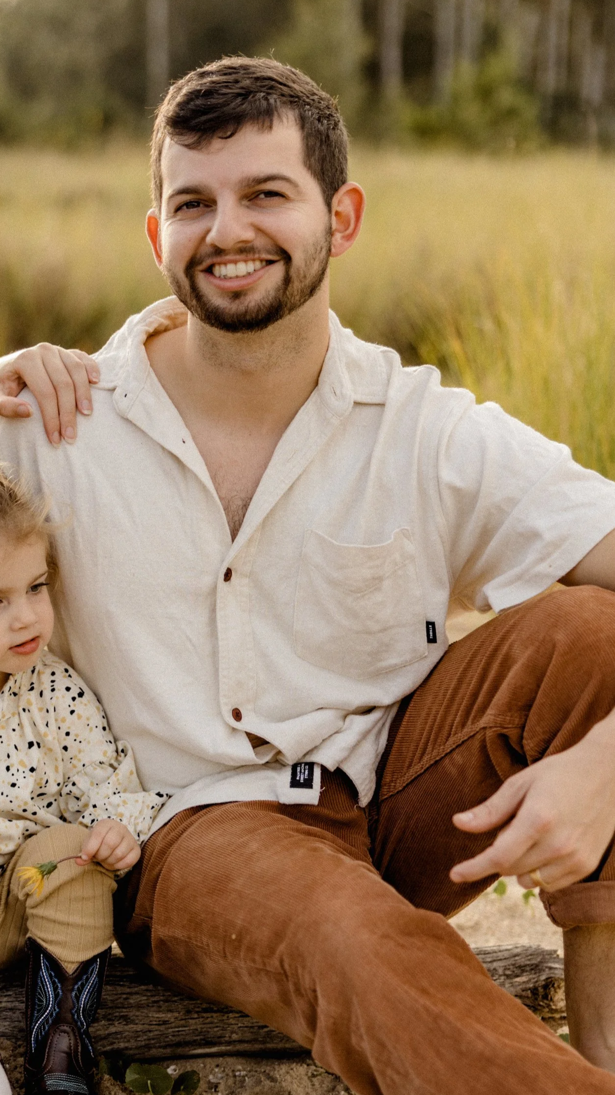 A man with short dark hair, beard, and mustache, smiling, sitting outdoors in a natural setting.
