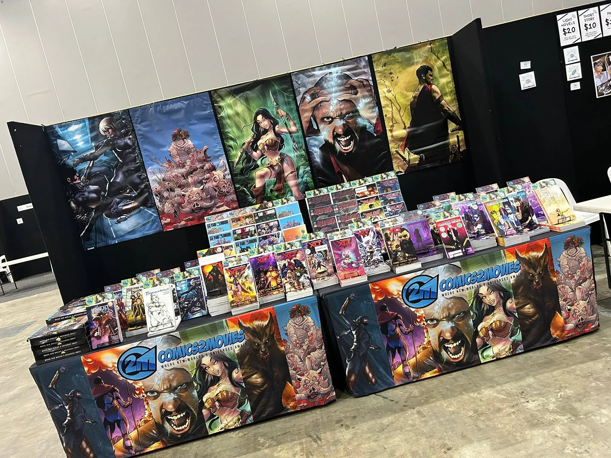 Display booth at a comic convention with large colorful posters of fantasy characters, manga, and graphic novels, along with various comic books and merchandise arranged on tables.