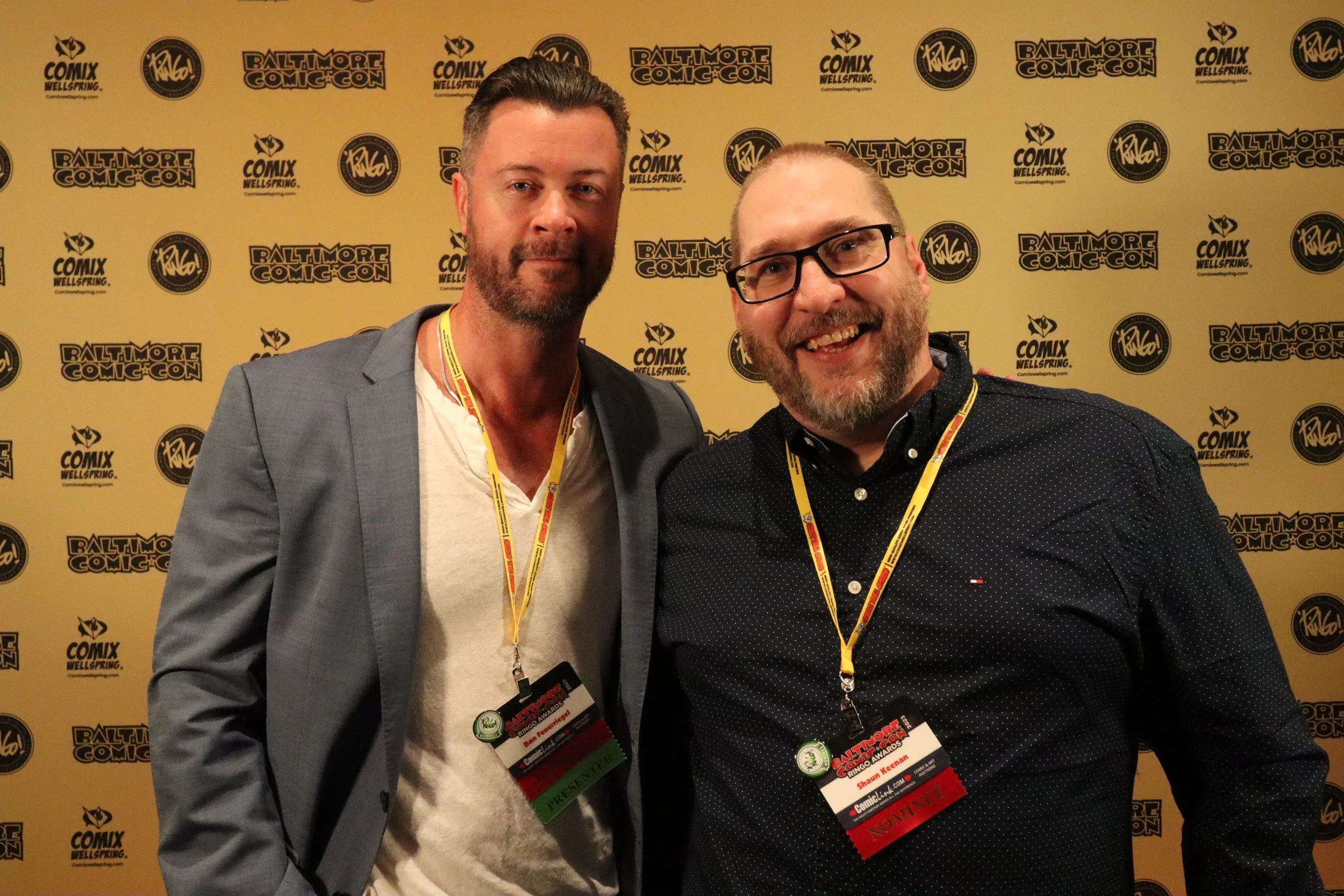 Two men posing at Baltimore Comic-Con with a yellow background featuring comic convention logos. Both men are wearing lanyards and badges; one has glasses and a beard, the other has short hair and a beard.