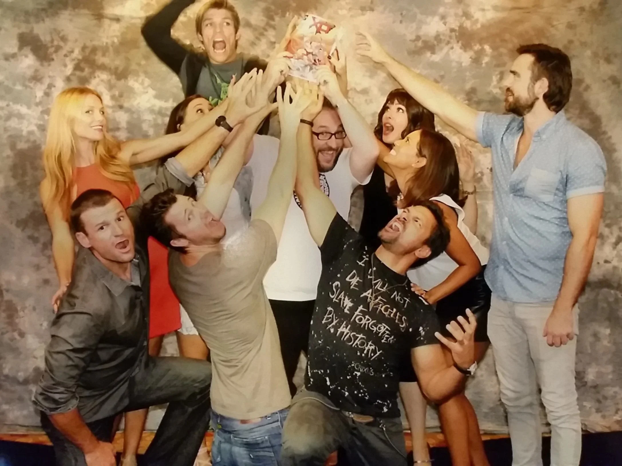 A group of people at a celebration or party, cheerfully reaching up together towards a small, colorful, novelty cake or object, with a textured, neutral backdrop.