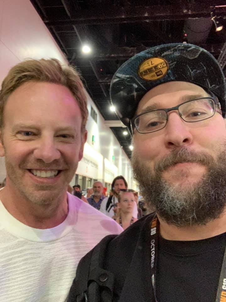 Two men smiling for a selfie at an indoor event with a crowd in the background.