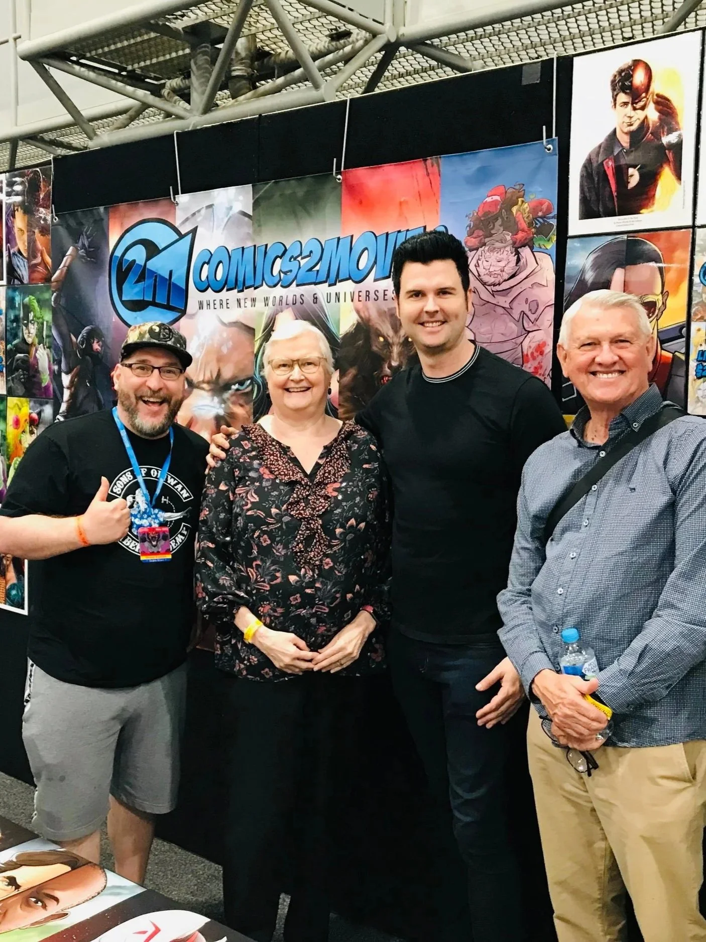Four people standing in front of a booth with a banner that says "COMICS2MOVIES." The banner has colorful images of various comic book characters and artwork. The group appears happy and are smiling, with some giving thumbs-up signs.