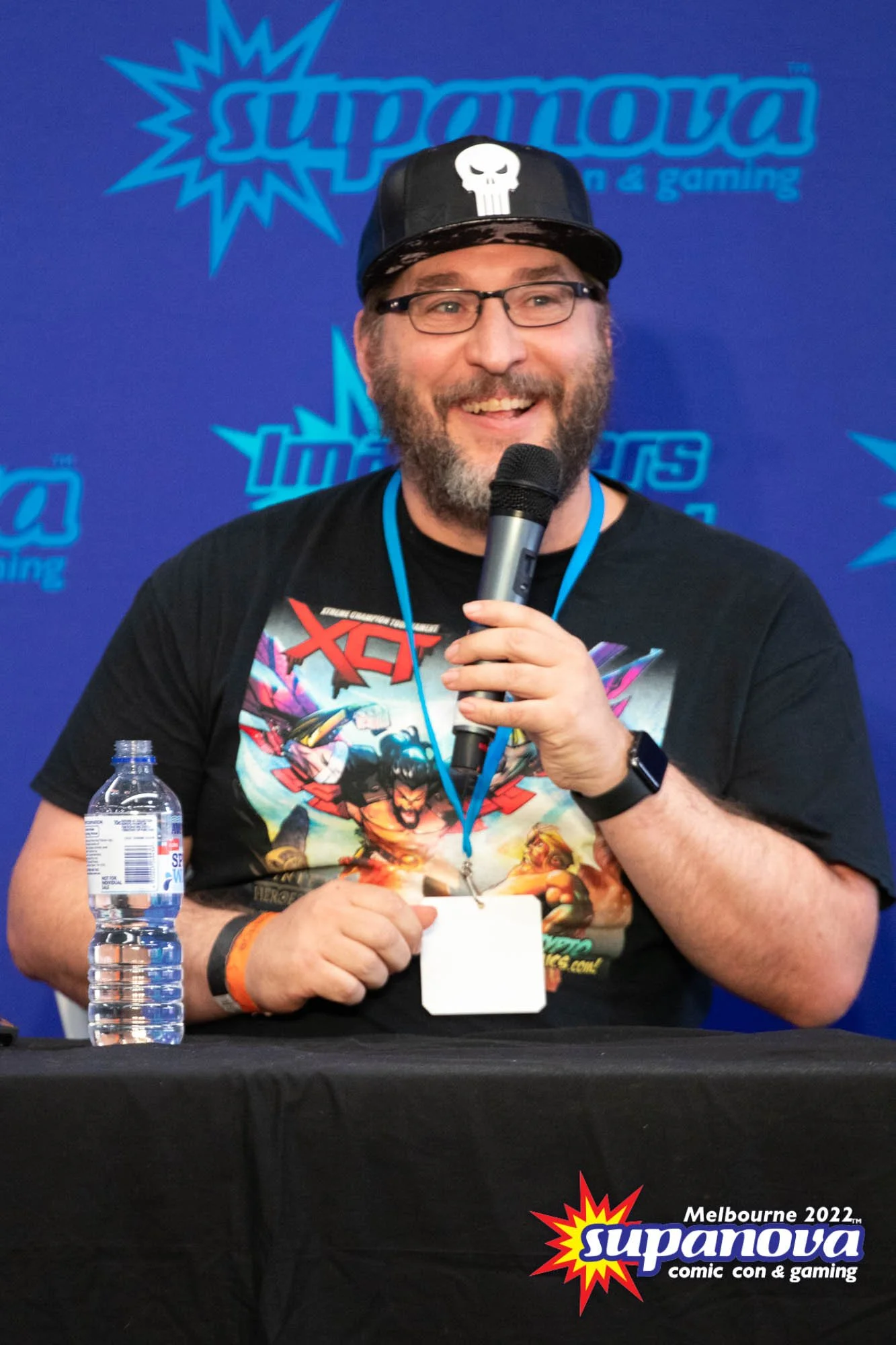 A man with glasses, a beard, and a cap with a white skull logo, smiling and holding a microphone at a comic convention. He wears a black T-shirt featuring comic book characters. A water bottle is on the table in front of him. Behind him is a blue backdrop with the 'Supanova' logo, and the text 'Melbourne 2022,' indicating the event location and year.