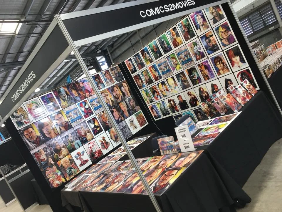 A booth at a convention displaying comic book and movie art prints of various animated and comic characters on a wall and table, with a sign showing prices.