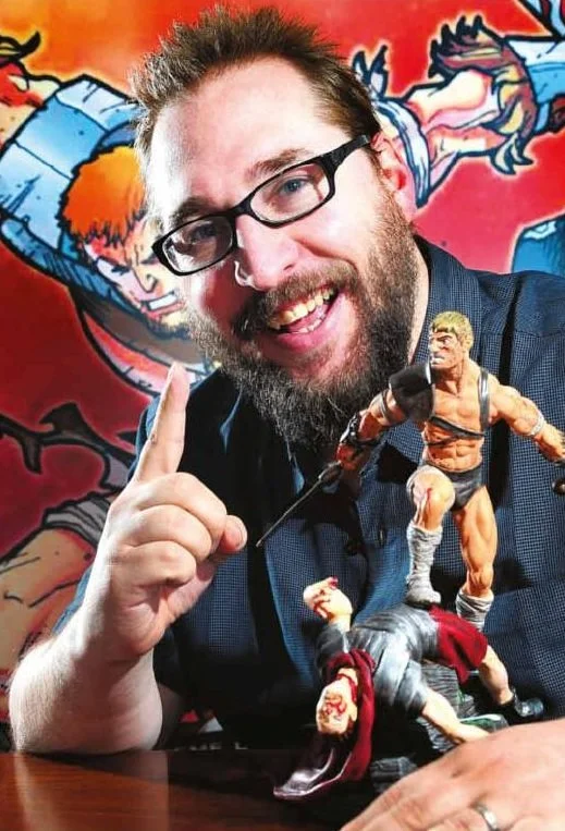 Man with glasses and beard smiling, pointing upward, sitting at a table with action figures, with colorful cartoon background.