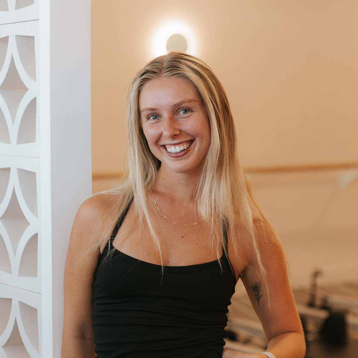 A young woman with long blonde hair smiling, wearing a black tank top and a gold necklace, standing indoors against a beige wall with a white decorative partition on her left.
