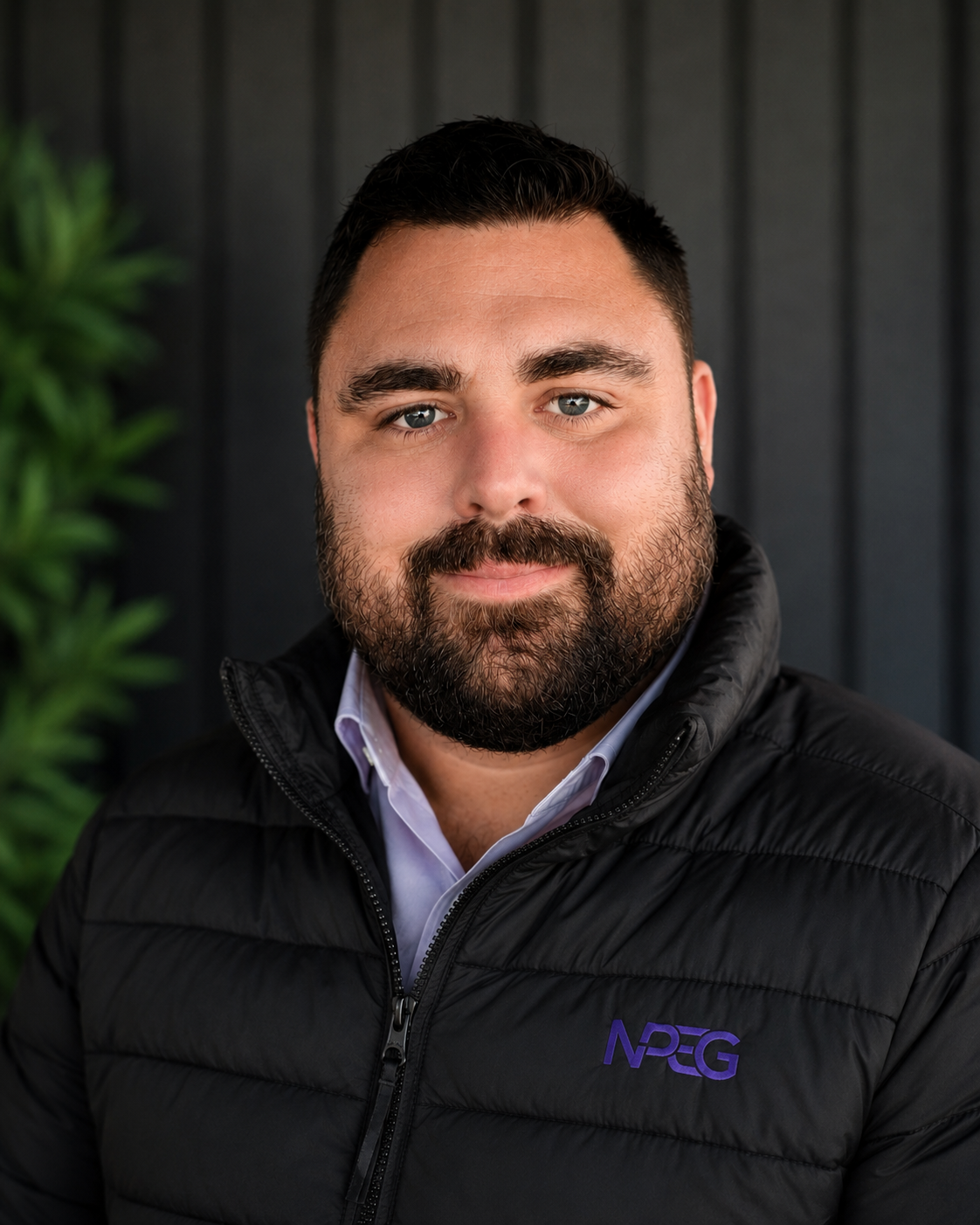 Portrait of a man with dark hair and a beard, wearing a black puffer jacket with a purple logo, standing in front of a dark background with some greenery.