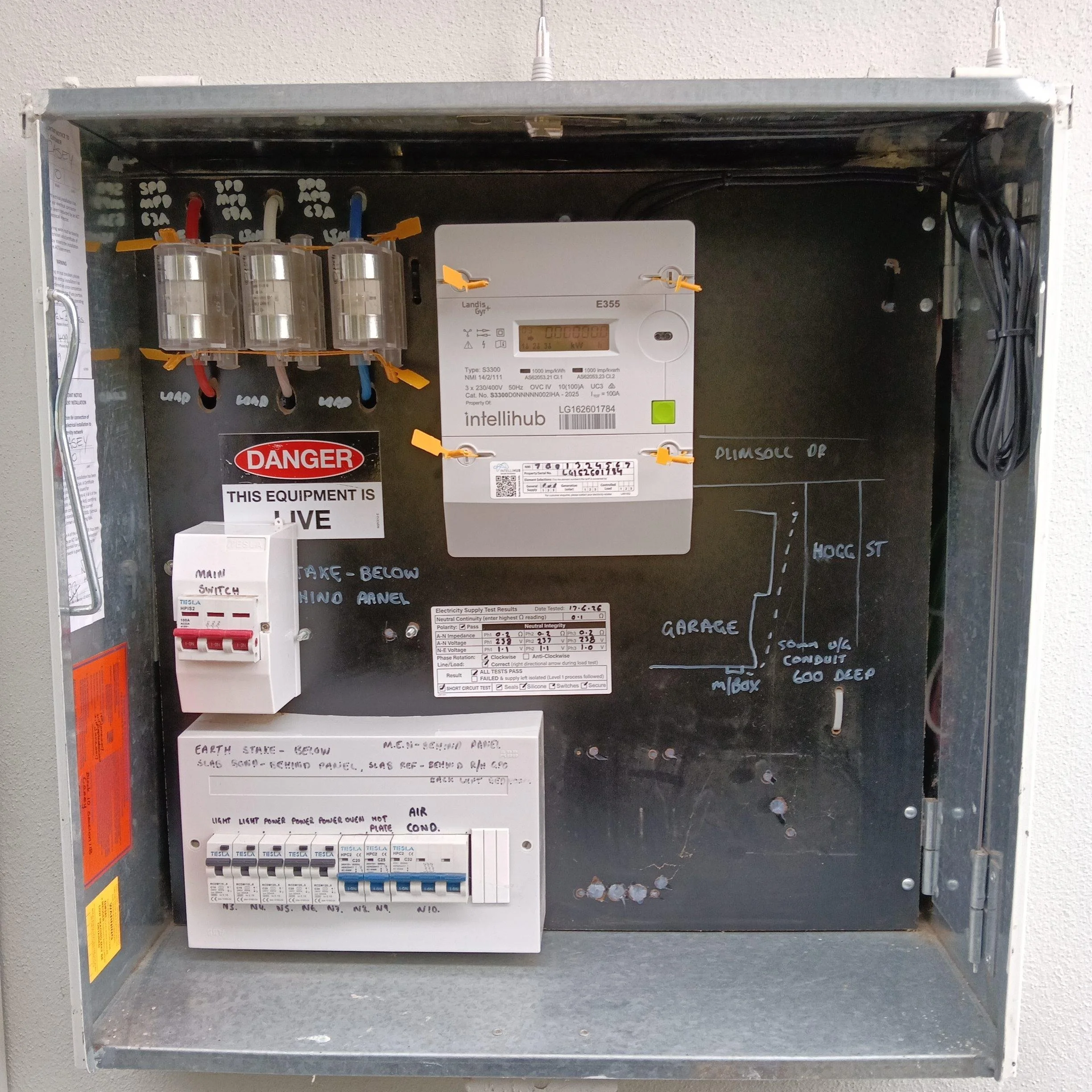 Open electrical panel with circuit breakers, wiring, and a digital meter, mounted on a wall with handwritten notes and safety warning stickers.