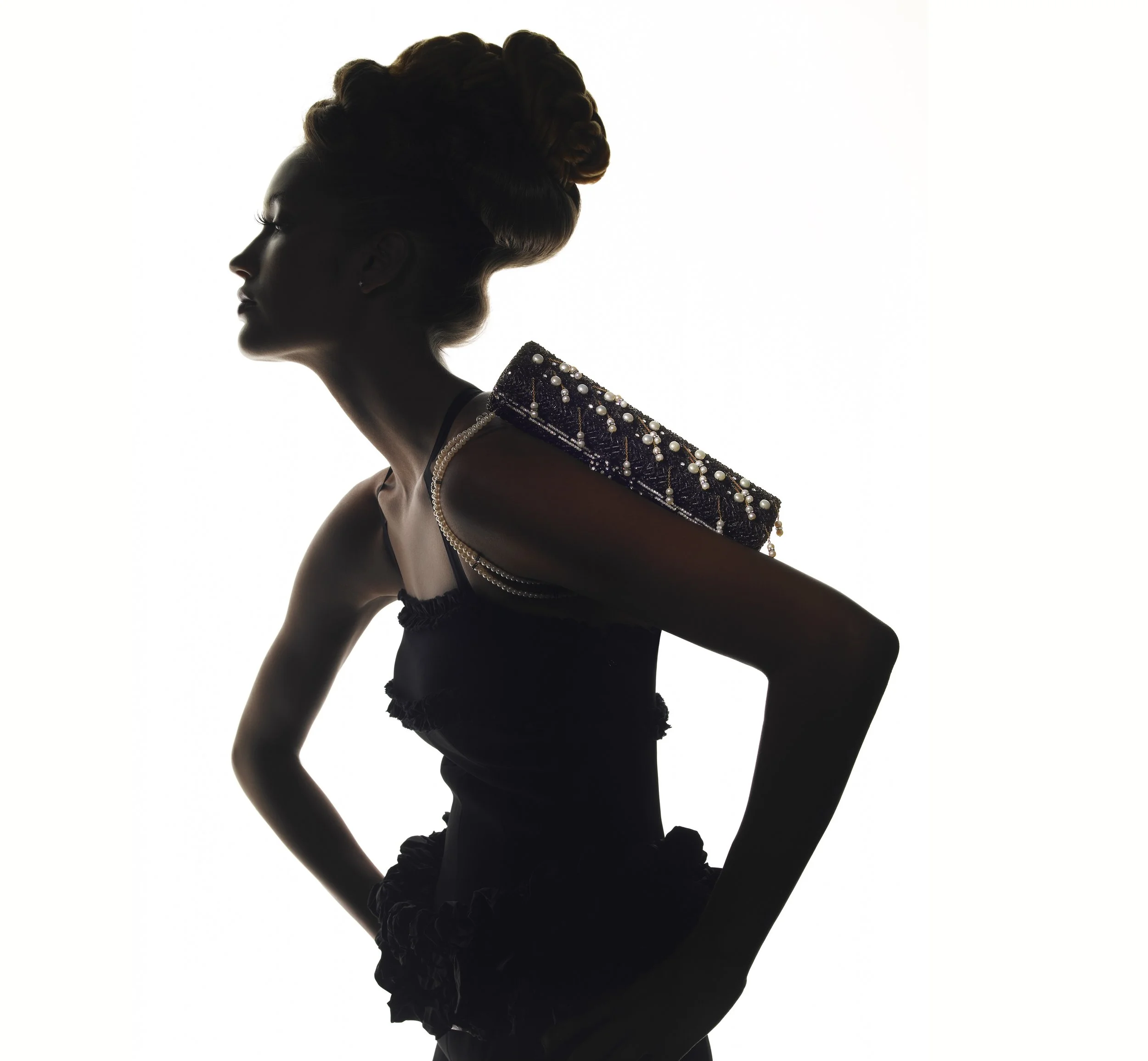 Silhouette of a woman with an elaborate hairstyle and adorned with pearl necklaces and a bejeweled shoulder piece, set against a white background.
