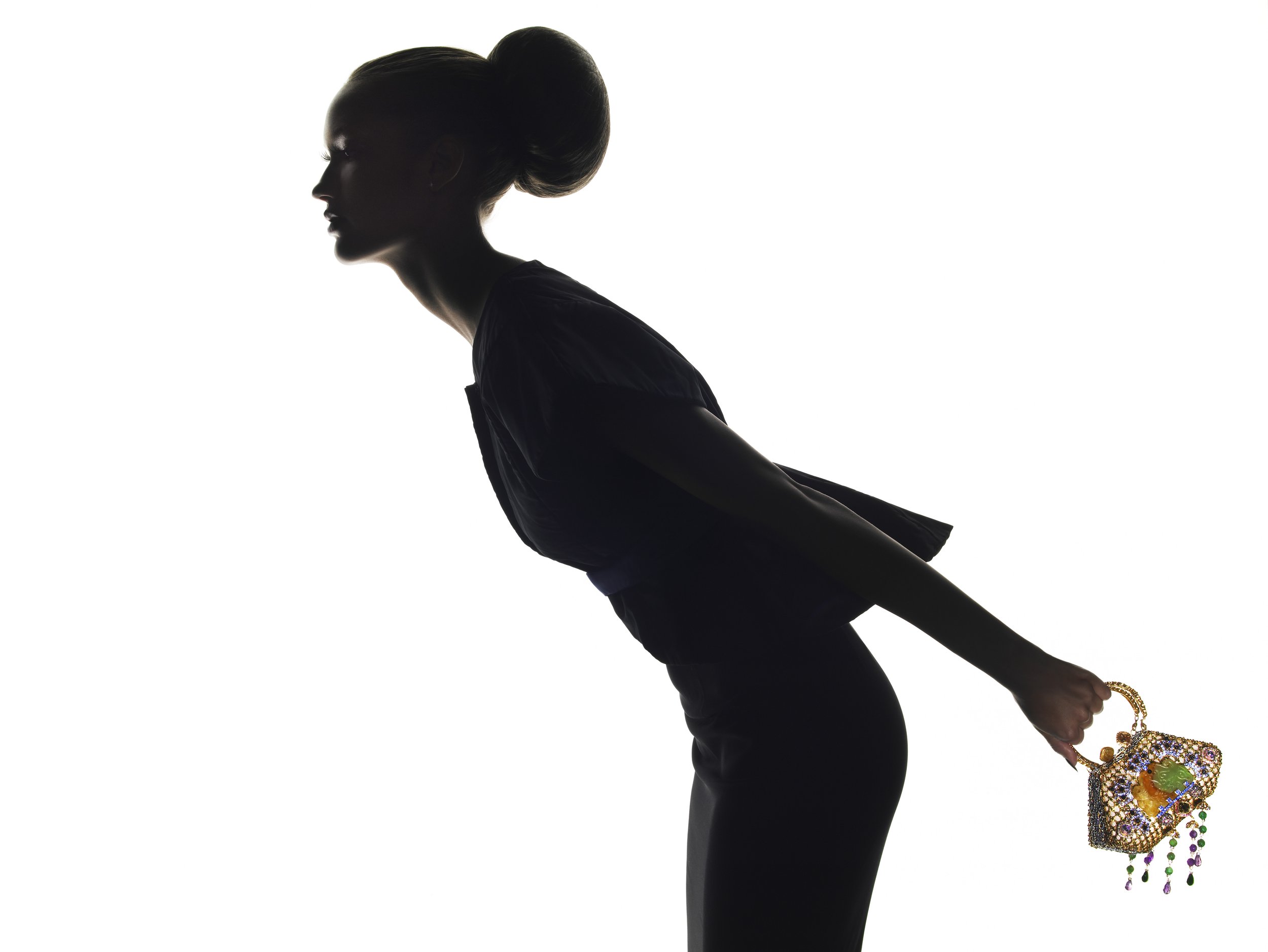 Silhouette of a woman with her hair in a big bun, kneeling and leaning forward, holding a colorful beaded handbag with jewelry.