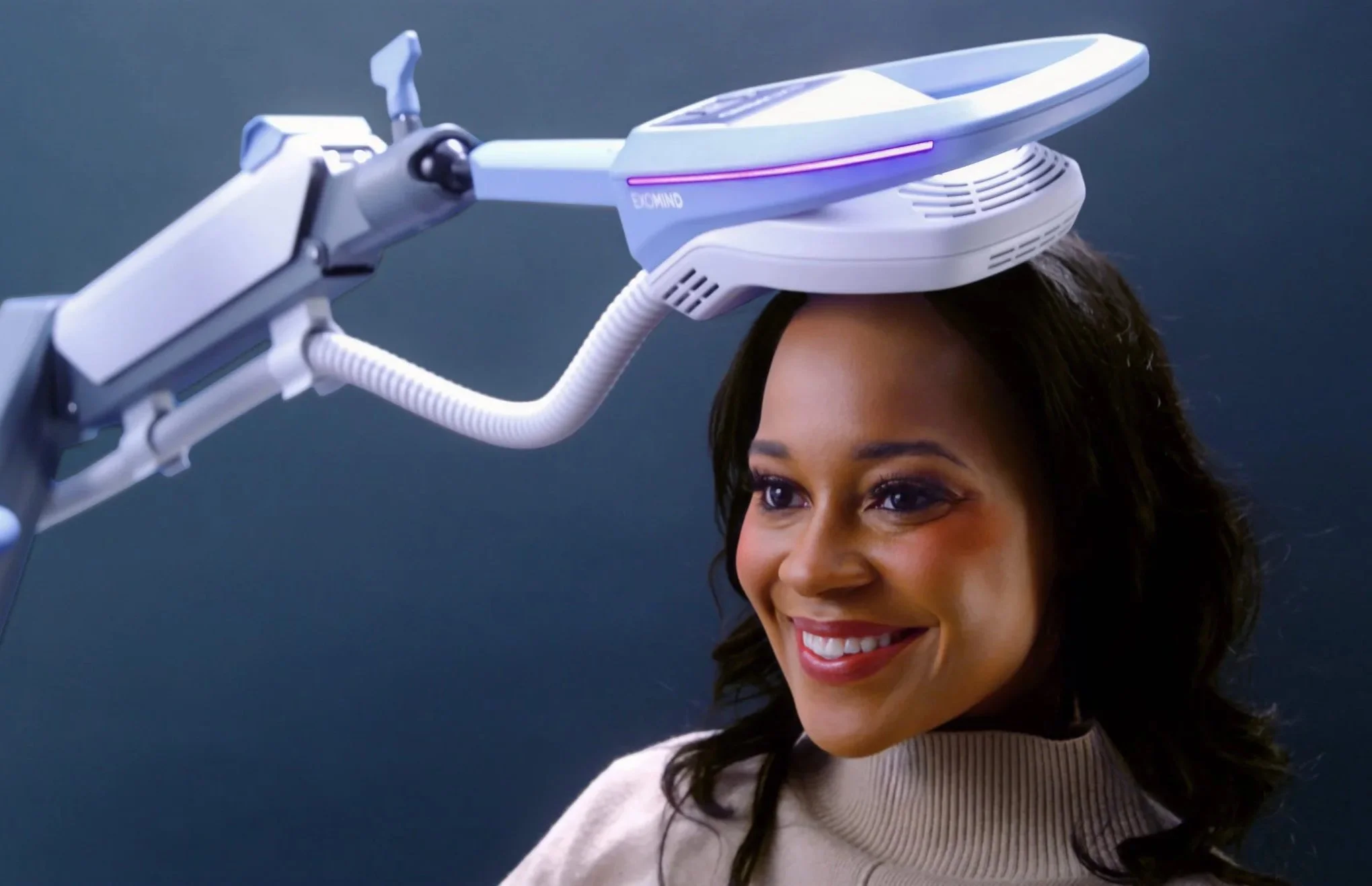 A smiling woman with long dark hair wearing a beige sweater gets a futuristic headband or helmet placed on her head by a robotic arm.