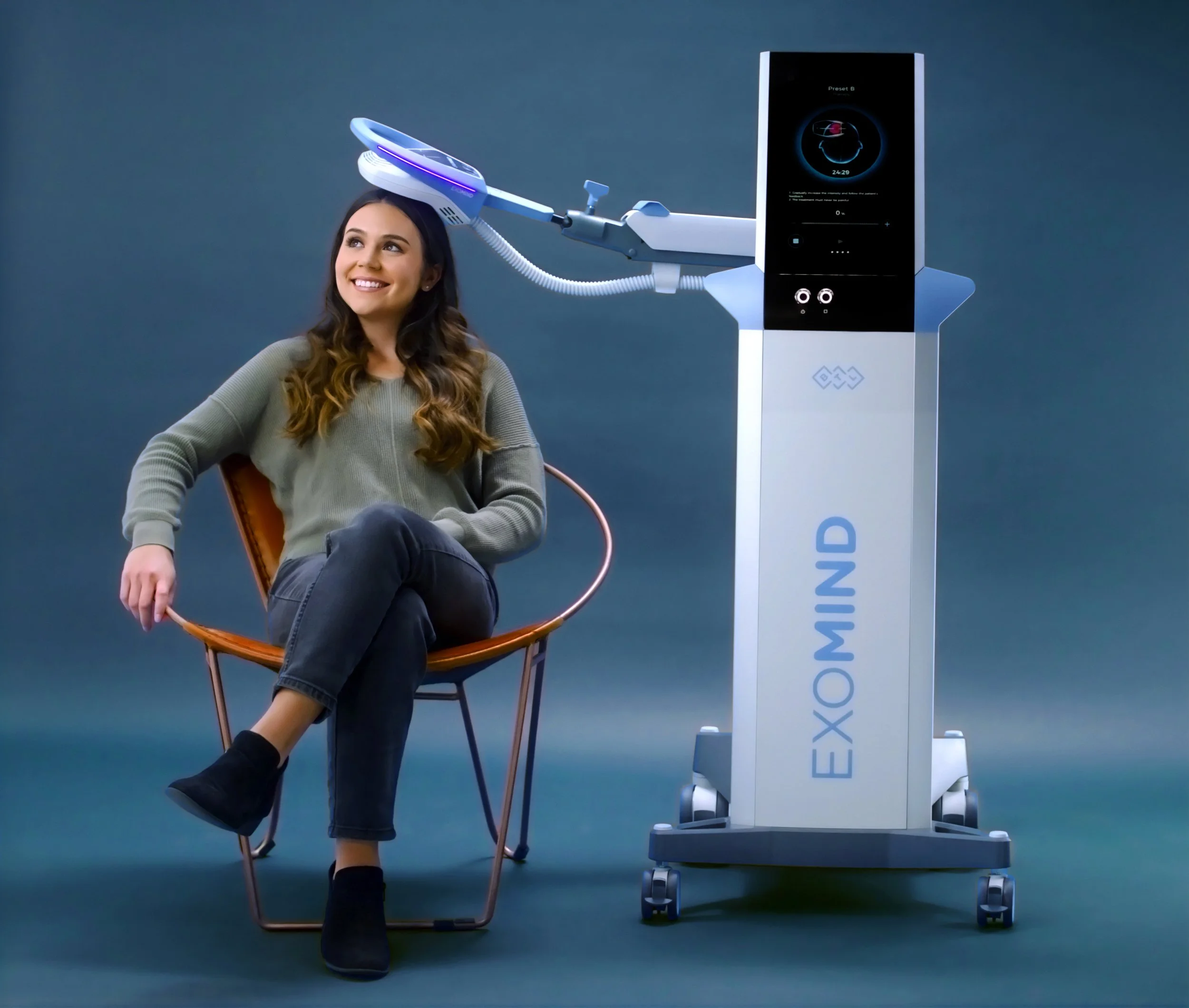 A young woman with long dark hair sitting on a chair, smiling, wearing a gray sweater and black boots, with medical or cosmetic equipment next to her, including a large machine with a screen and robotic arm.