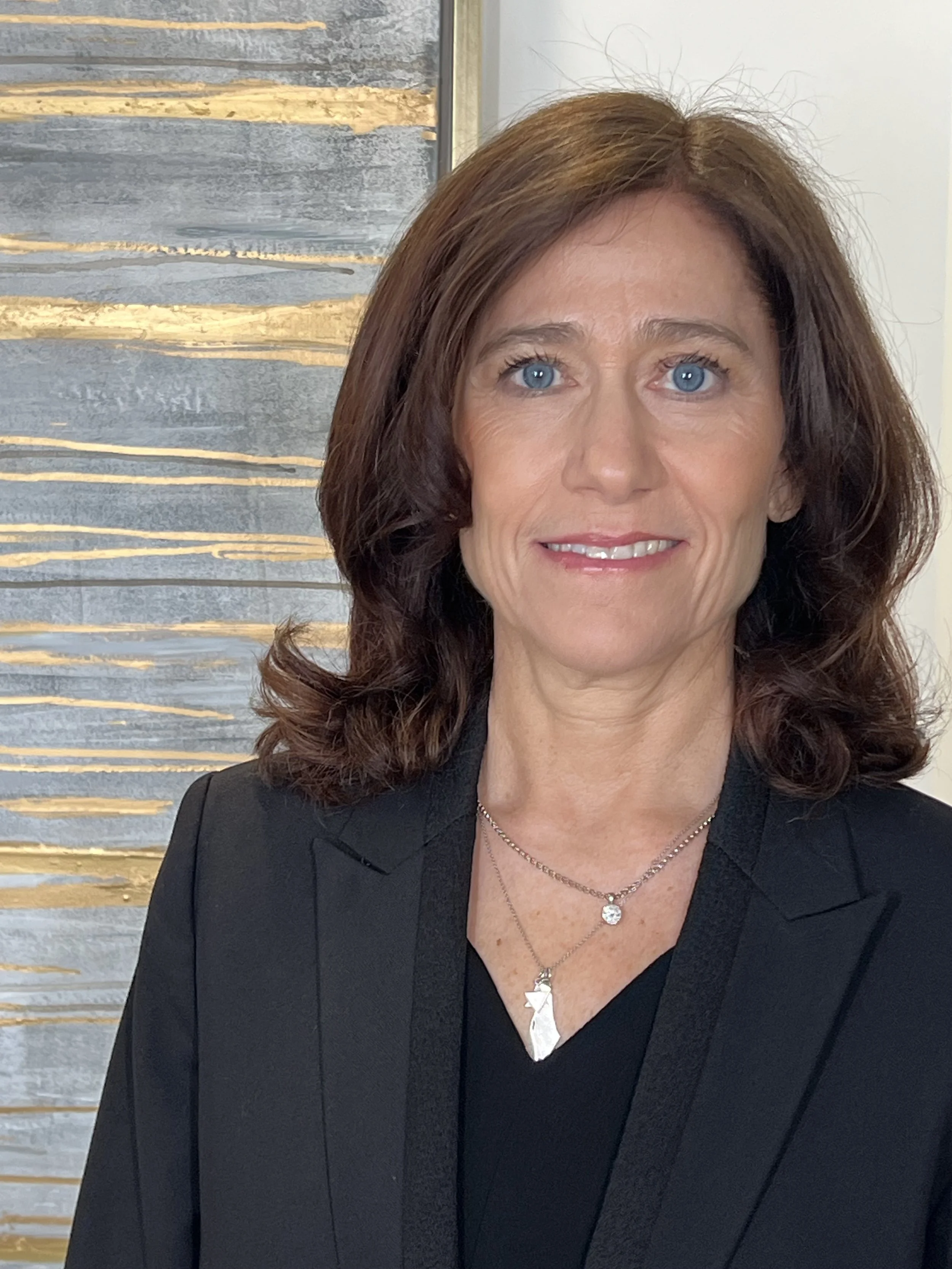 A woman with shoulder-length brown hair and blue eyes, smiling, wearing a black blazer and layered necklaces, standing in front of an abstract background with gold and grey streaks.