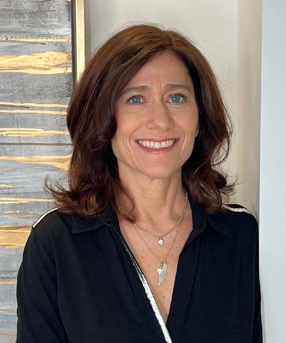 A woman with shoulder-length brown hair and blue eyes, smiling, wearing a black top and layered necklaces, standing indoors near a modern art piece.