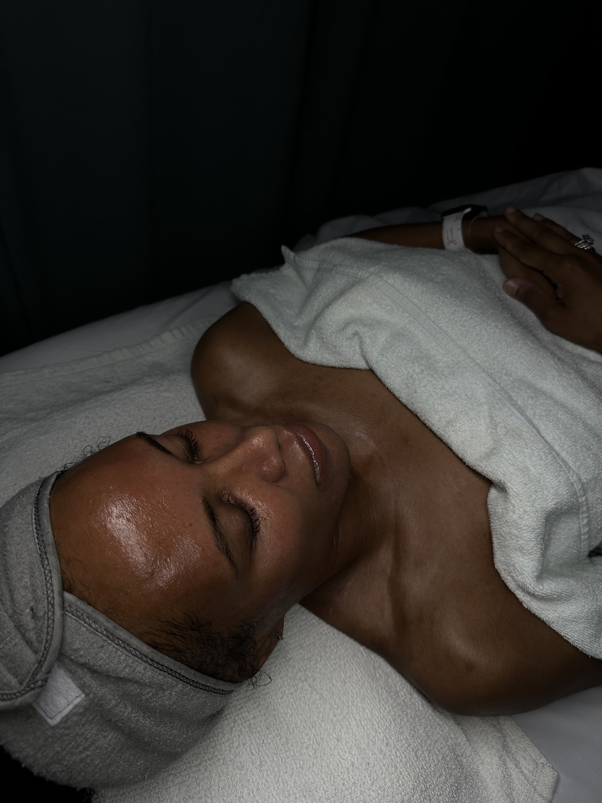 A woman lying on a bed with a towel draped over her chest, wearing a gray cap and a ring, with her eyes closed, appearing to be receiving a massage or facial treatment in a spa or clinic setting.