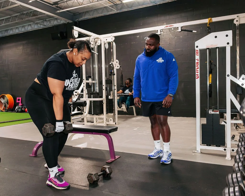 A client lifting dumbbells while founder, Micah Baisden, in a blue jacket with the PowerHouse Sports Academy logo observes and provides guidance in the gym.