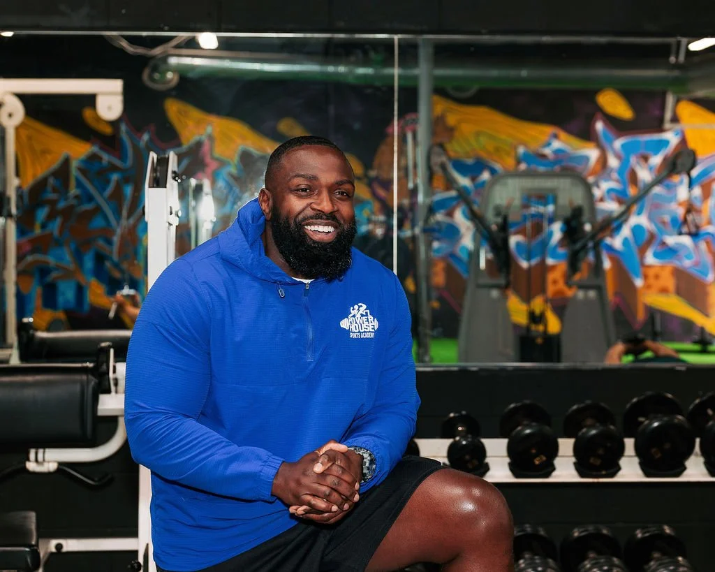 Founder, Micah Baisden, smiling in the gym with colorful graffiti on the wall behind him, wearing a blue PowerHouse Sports Academy jacket and black shorts, seated in front of dumbbells.
