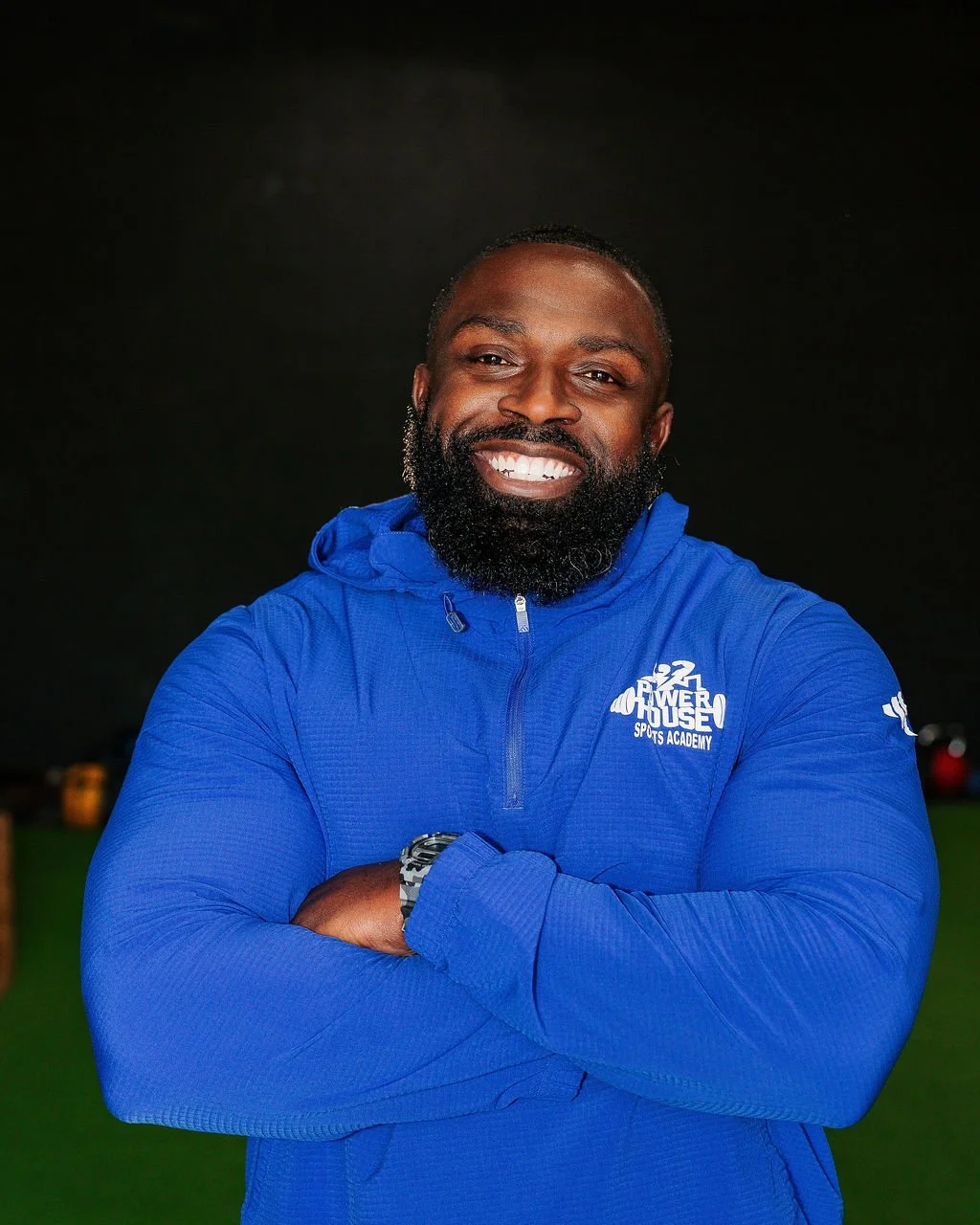 Micah Baisden, the founder, smiling and wearing a blue athletic jacket with 'Power House Sports Academy' logo, standing with crossed arms against a dark background.