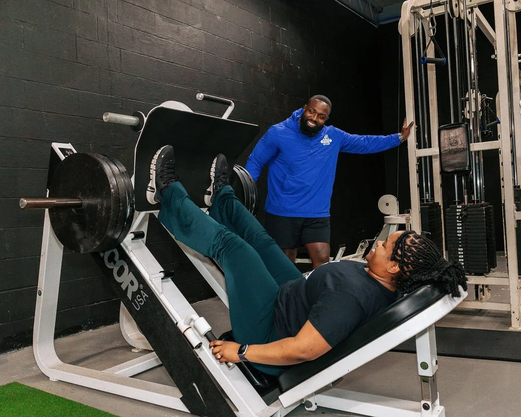 A client is performing a leg press exercise in the gym, lying on a leg press machine with the founder, Micah Baisden, standing nearby, smiling and holding the machine. The gym has black walls and is equipped with various workout machines.